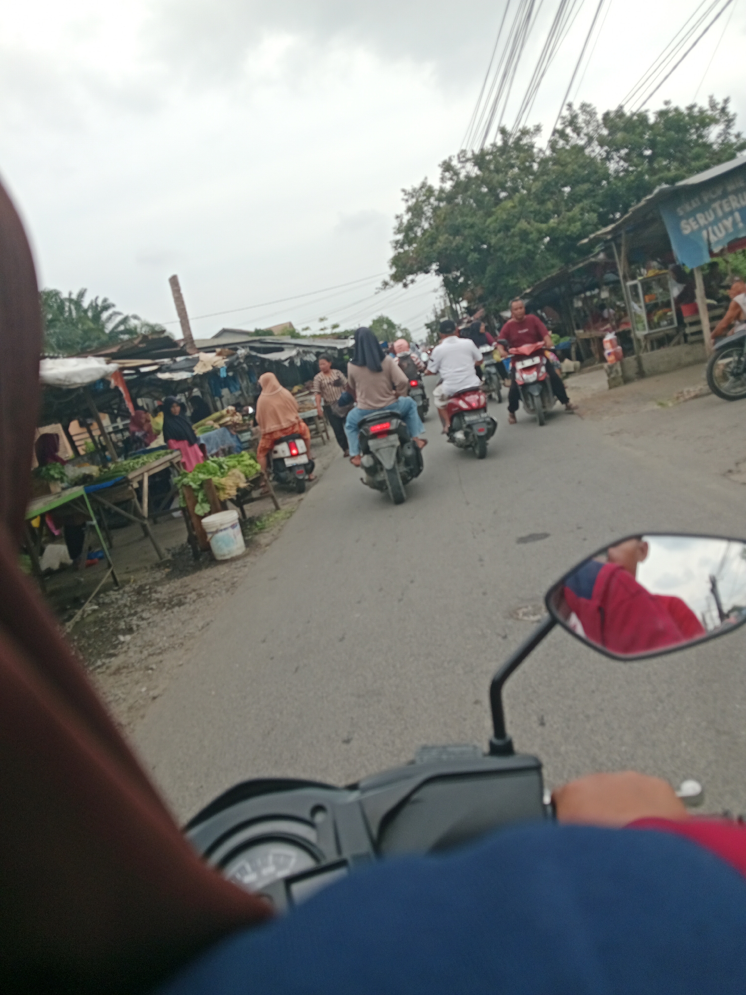 The image shows a busy street scene, likely in the city of Binjai, Indonesia, with several motorcycles and vendors. The perspective is from someone riding a motorcycle, looking forward at the road and the activity. On the right side of the road, there are market stalls with produce, and people are seen shopping or working. A blue sign with white lettering is partially visible on one of the stalls. Several people are riding motorcycles, some carrying passengers. The sky is overcast. The foreground shows a portion of the motorcycle the viewer is on, including the handlebars and a rearview mirror reflecting a person in a red shirt.