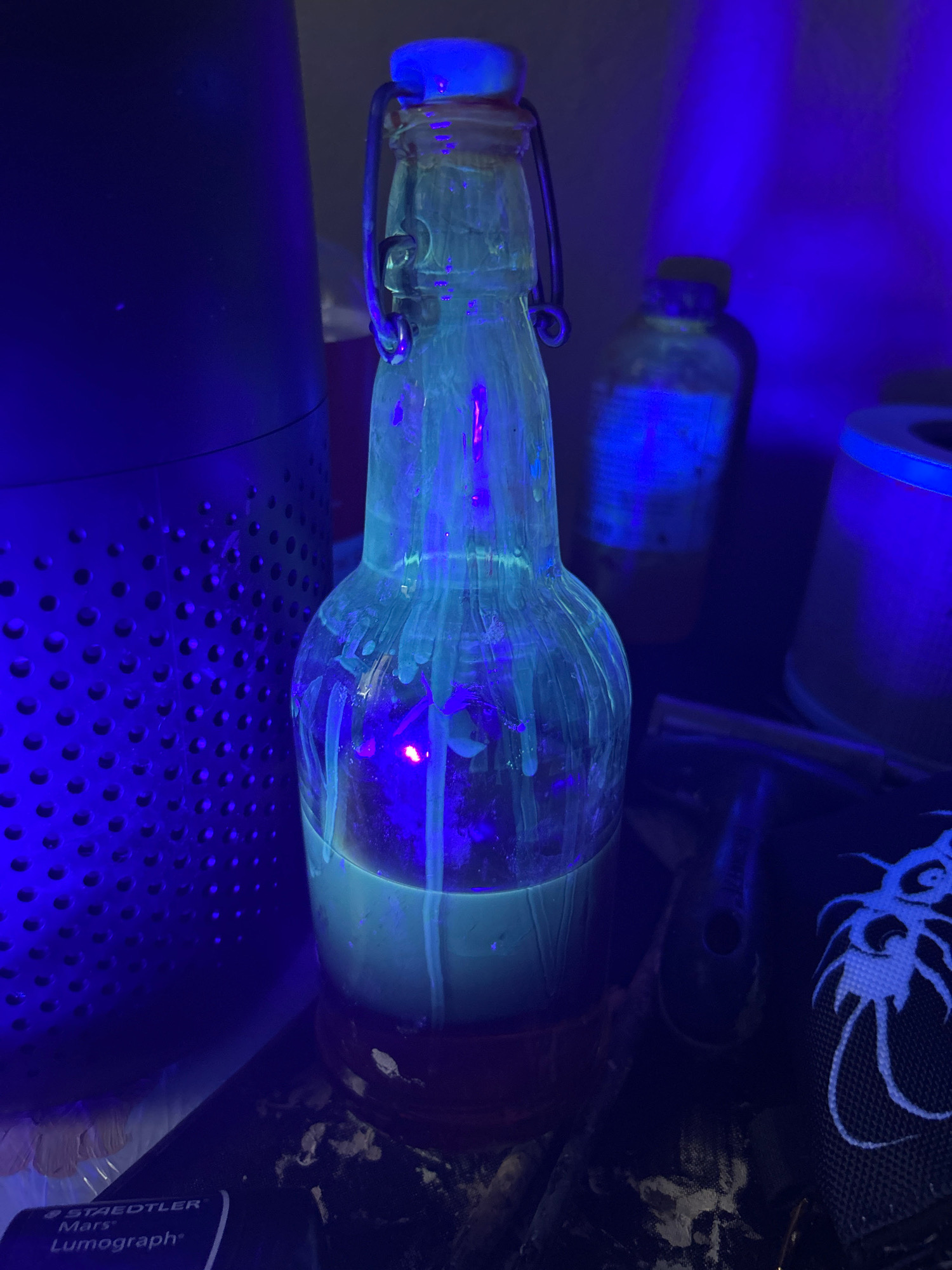 A glass bottle with a flip-top lid, filled with a pale liquid and illuminated by a black light, sits on a dark surface. The liquid inside the bottle glows a bright blue-green, and streaks of the same glowing substance drip down the sides of the bottle. Behind the bottle, a dark, perforated cylinder is visible, also under the black light, emitting a strong blue glow. To the right, another bottle with a yellow cap is visible, with a faint blue glow emanating from it. In the foreground, a black object with a white, web-like design is partially visible. The scene is dark overall, with the black light creating a vivid, artificial illumination. The location is not specified in the image or its metadata.