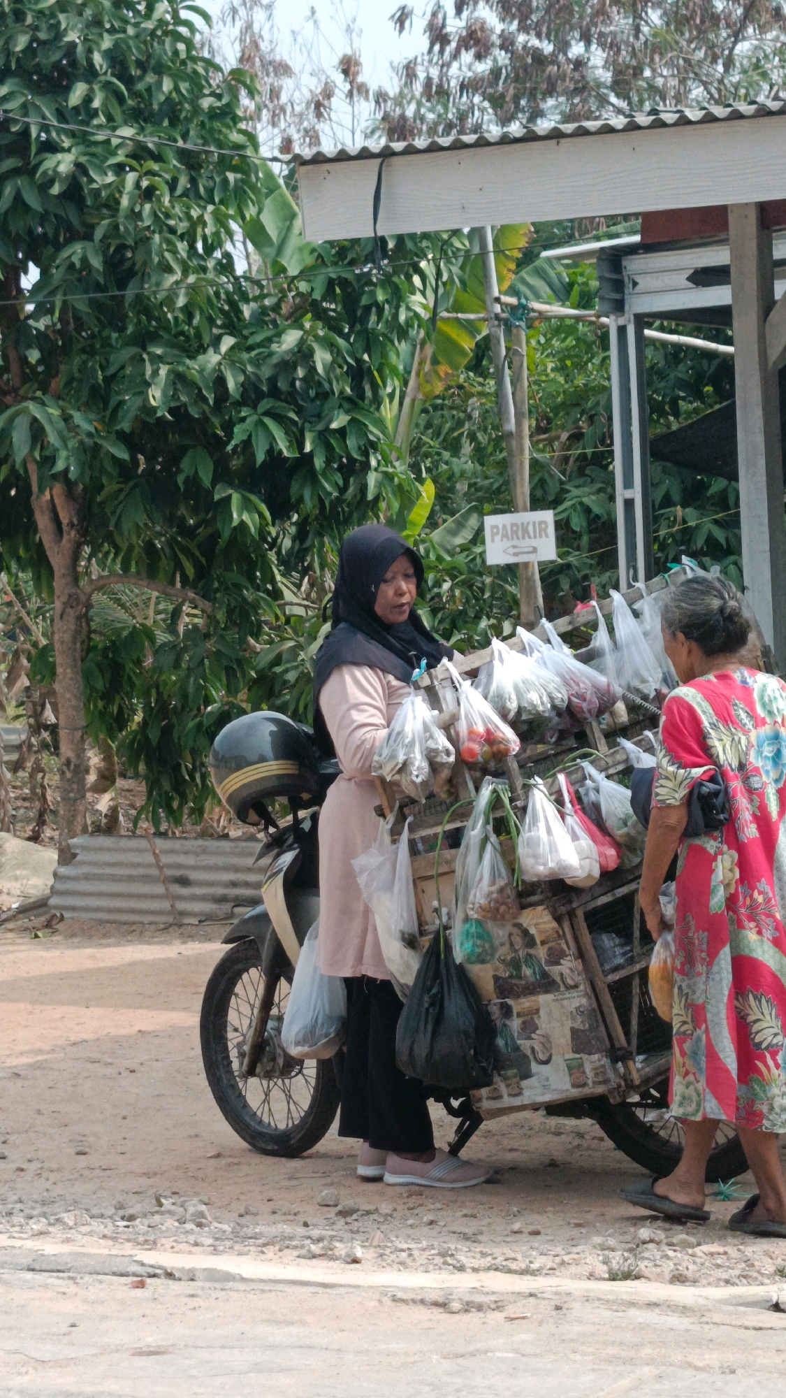 In Metro, Indonesia, two women are engaged in a transaction at a roadside market. A woman wearing a black hijab and a light pink abaya stands next to a motorcycle, holding a variety of plastic-bagged produce. She appears to be the vendor. In front of her, a woman in a vibrant, floral-patterned dress faces the vendor, potentially as a customer. Their interaction is focused on the goods displayed on a wooden cart attached to a motorcycle. The environment suggests a warm, sunny day with lush greenery and a corrugated metal roof overhead. A sign that reads "PARKIR" with an arrow indicates a parking area, suggesting the location is a public space. The ground is dusty and uneven, typical of a street or outdoor market setting.