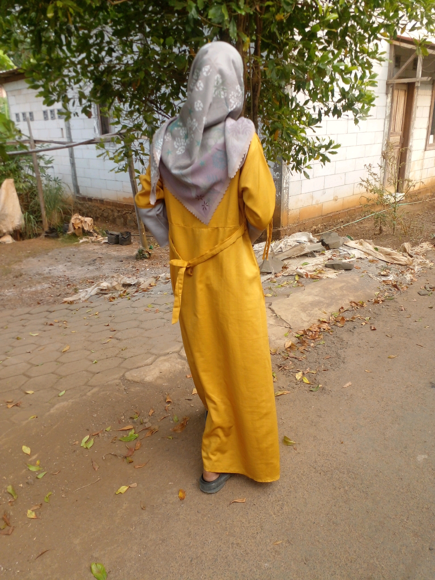A person wearing a yellow dress and a grey hijab stands with their back to the camera on a paved surface in Pungangan, Indonesia. They are standing on a path that appears to be made of concrete pavers, with loose dirt and fallen leaves scattered around it. Behind the person is a white brick building with a wooden door and window. To the left of the building, there is a tree with green leaves. The ground in front of the building is also covered with dirt and debris, including some white fabric and grey stones. The overall scene suggests an outdoor environment, possibly a residential area or a street in a village.
