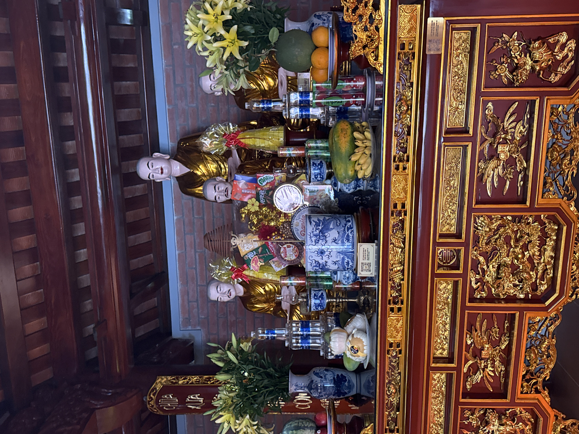 The image shows a Buddhist altar at the pagoda of Diên Khánh, Vietnam. The altar is adorned with offerings, including fruit, flowers, and drinks. Several statues of Buddha are displayed prominently on the altar, along with intricate blue and white porcelain vases. The altar itself is a richly decorated wooden structure with ornate golden carvings. The background features a brick wall and a wooden ceiling with exposed beams, typical of traditional Vietnamese architecture.