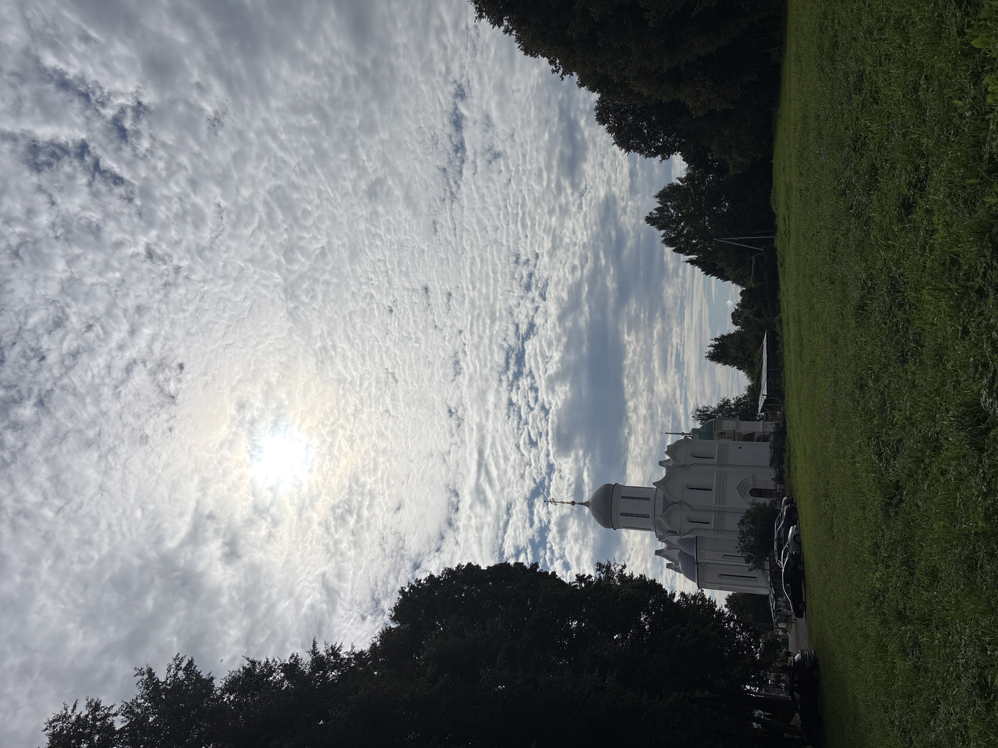 A white church with a grey dome and cross stands on a grassy hill under a cloudy sky. The church has multiple arched windows and decorative architectural elements. Several cars are parked in front of the church, and a few trees are visible on the right side of the image. The sky is filled with white, puffy clouds, with the sun peeking through in the upper center. The foreground is a green, mowed lawn. This is the Savvino-Storozhevsky Monastery in Zvenigorod, Russia.