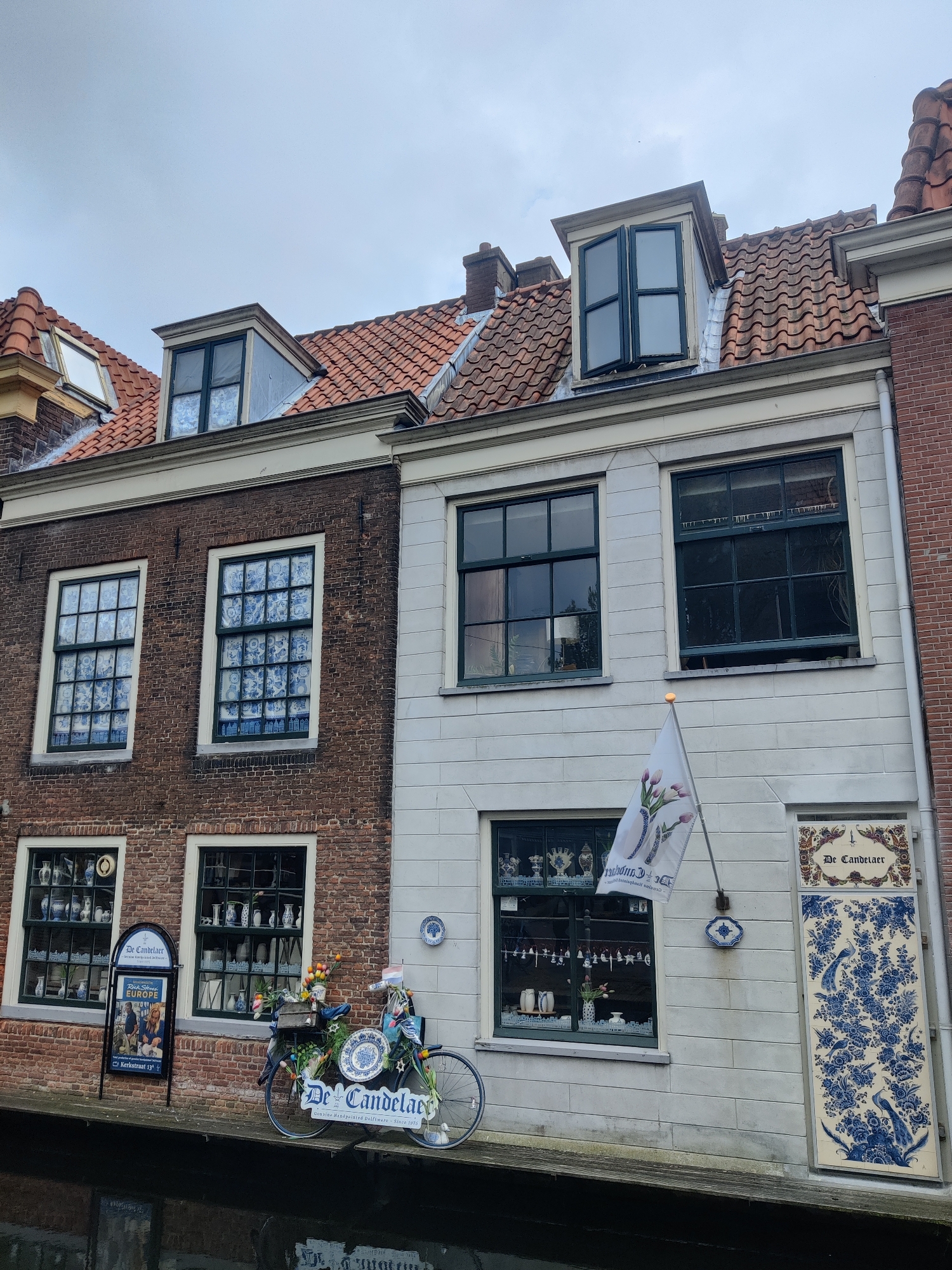 The image depicts a Dutch canal in Delft, Netherlands, with two brick buildings lining the waterway. The building on the left is made of dark red brick and features multiple windows with blue and white patterned curtains visible through them. The building on the right is constructed of white stone and has a large, decorative tile panel on its facade with blue floral and bird motifs, and the text "De Candelaer" above it.
In front of the white building, a bicycle is parked, adorned with a variety of decorative blue and white plates, flowers, and a flag. A signpost with information about "De Candelaer" and an advertisement for "Europe" stands near the bicycle. The windows of both buildings display porcelain and ceramic ware, suggesting that they might be shops or workshops. The canal water reflects the overcast sky and the architecture. The overall atmosphere is quaint and historical, characteristic of Delft's well-preserved cityscape.