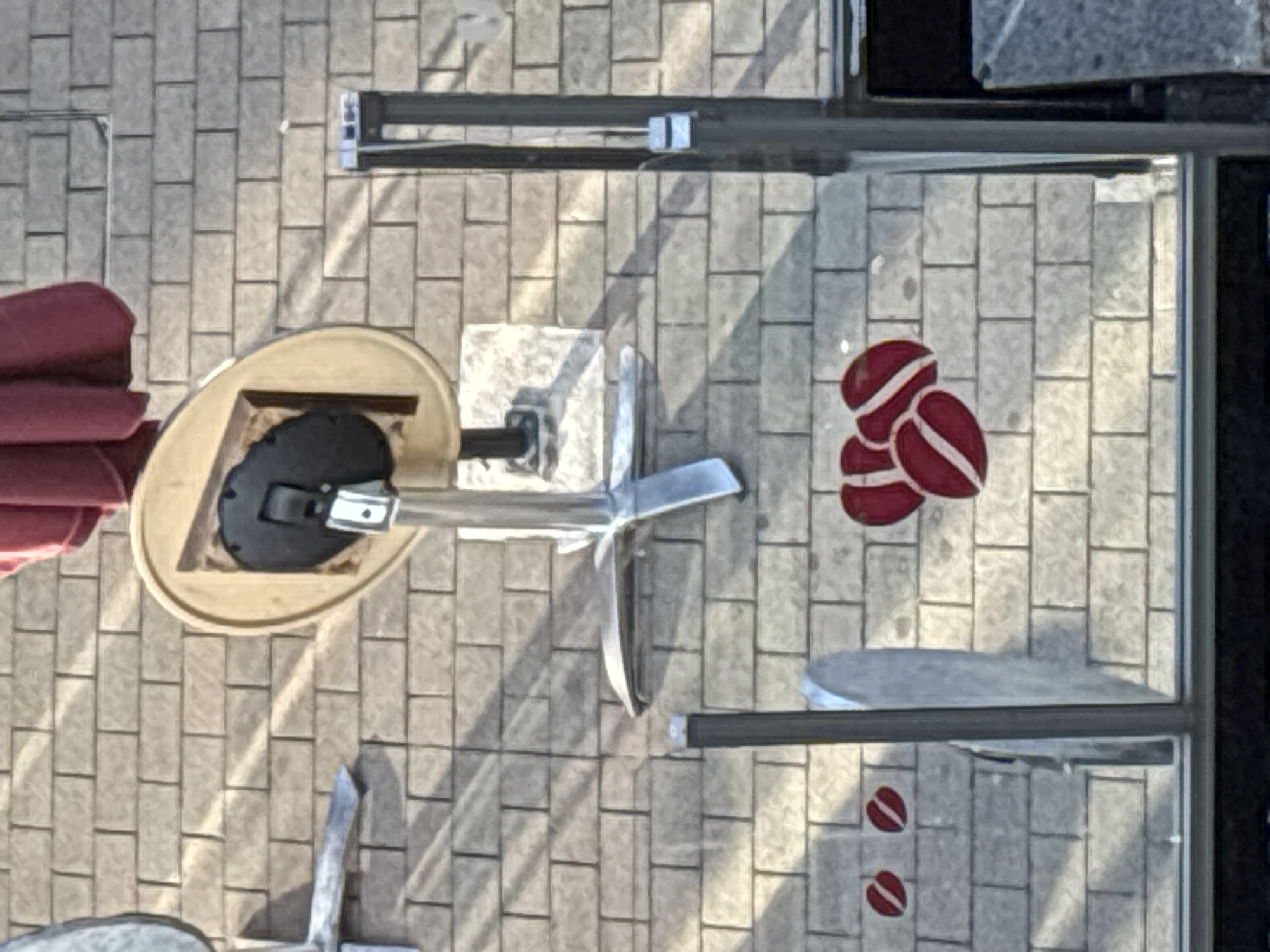 The image, taken from an elevated vantage point, shows an outdoor cafe setting with a brick-patterned tiled floor. In the center, a table with a round, light-colored top and a dark base is partially visible, with a red umbrella positioned above it. To the left of the table, a section of a metal table leg is seen.
In the foreground, through what appears to be a glass barrier, a brick-patterned wall is depicted. On this wall, there are decorative elements resembling coffee beans in a deep red color with white stripes. Two such decorative elements are positioned to the left, and a cluster of three is to the right. Vertical gray metal posts frame the view of the wall and decorations. The lighting suggests it is daytime, with shadows cast across the brickwork. The scene is devoid of any people.
While the image itself does not explicitly contain information about its location in Cork, Ireland, the presence of outdoor cafe furniture and the general aesthetic are consistent with urban environments. This type of setting is common in many city centers, including those in Ireland.