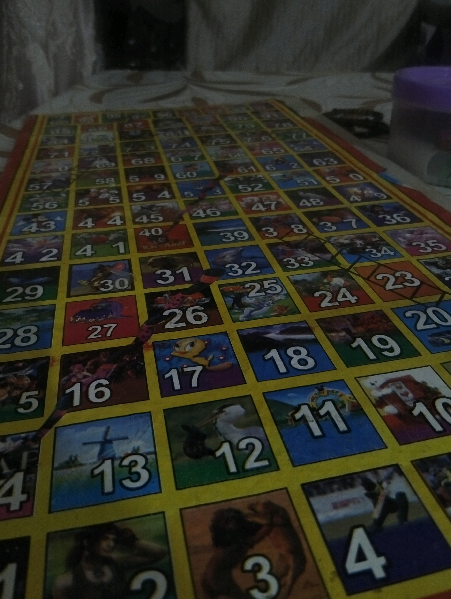 The image shows a close-up, low-angle shot of a Snakes and Ladders board game, laid out on a table. The game board is colorful, with squares numbered from 1 to 100, featuring various cartoon illustrations. Some squares have numbers partially obscured or are covered by what appears to be a torn section of the game. The lighting is dim, suggesting an indoor setting, possibly at night or in a poorly lit room. The background shows blurred fabric curtains and a dark, indistinct area, hinting at a residential or casual environment. No people are visible in the image. The board is oriented at an angle, with the lower left corner closer to the viewer. The specific location in Gujrat, Pakistan cannot be determined from the image alone, as there are no identifying landmarks or contextual clues.