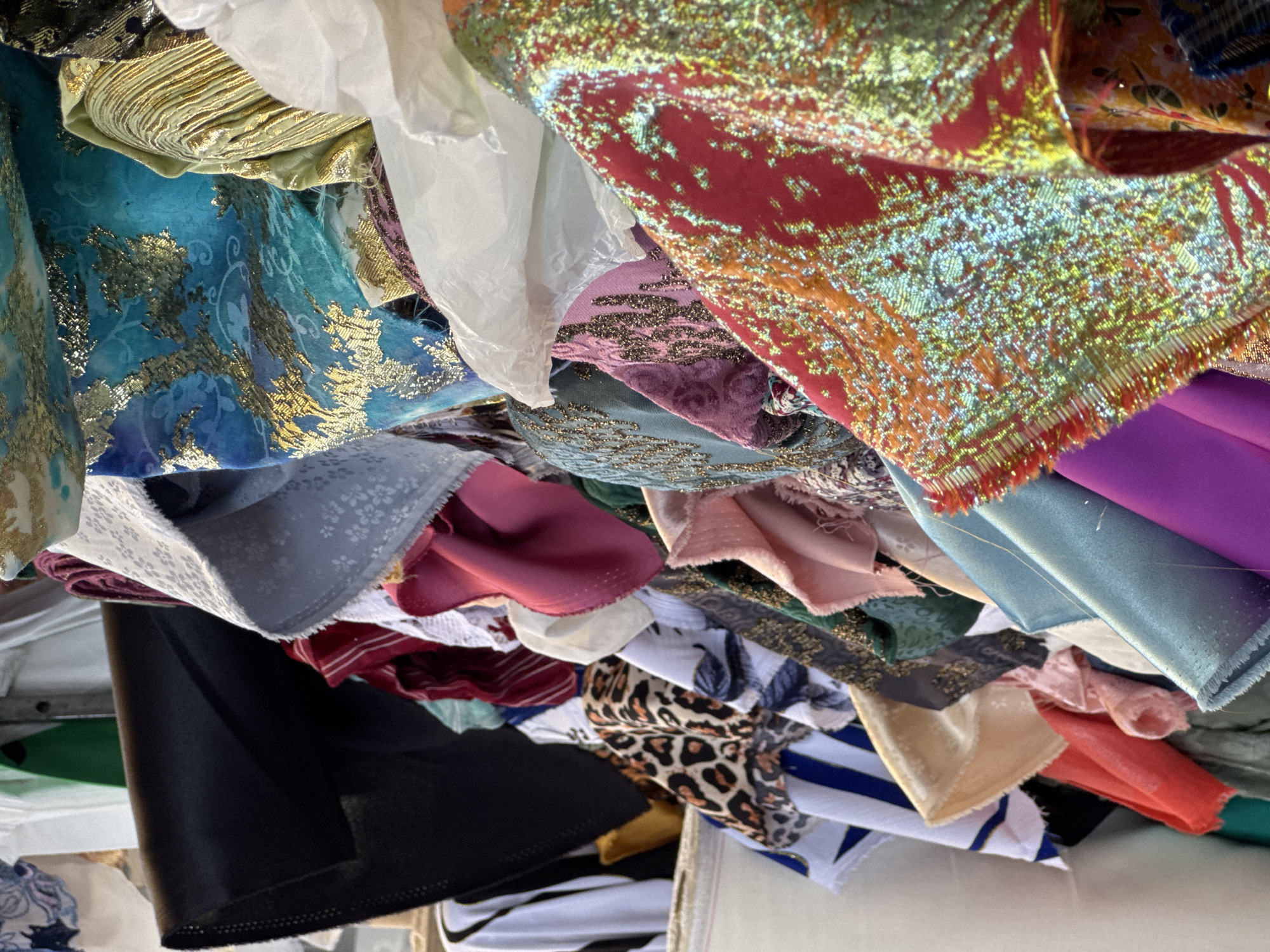 The image displays a chaotic, close-up, overhead view of a large pile of assorted fabrics and textiles. The composition is filled with a vibrant mix of colors, patterns, and textures, creating a dense and layered abstract scene.
**Subjects:** The primary subjects are numerous pieces of fabric.
**Objects:** The fabrics appear to be remnants, bolts, or lengths bundled together, possibly inside a container or piled on a surface. A crumpled white material, resembling tissue paper or a thin plastic bag, is visible among the fabrics.
**Scene/Setting:** The scene is an indoor setting, likely a fabric store, a sewing workshop, or a storage area for textiles. The fabrics are stacked or thrown together, suggesting either a state of disarray, an inventory, or a collection being sorted or stored.
**Activity or event:** No specific activity or event is taking place. The fabrics are static, implying a moment of storage or display rather than active use.
**Notable details:**
The fabrics showcase a rich variety:
* A prominent turquoise fabric with intricate gold floral and leaf patterns.
* A shimmering reddish-orange fabric with an iridescent, glitter-like surface of gold and green hues, and a fringed edge.
* A golden, pleated or ruched fabric.
* Solid colored fabrics in shades of purple, magenta, teal, black, and light green.
* Patterned fabrics including leopard print, white fabric with blue stripes, and a light grey fabric with subtle white floral or geometric motifs.
* Various textures are evident, from smooth satin-like finishes to heavily embroidered or sequined surfaces and crinkled materials.
* The lighting is diffused, typical of an indoor environment without direct sunlight.
* There is no visible text.
* No specific location cues are present within the image itself.