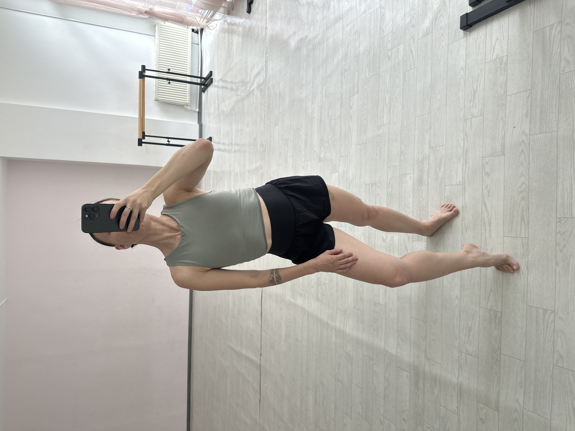 The image shows a full-body mirror selfie of a woman posing in a dance studio. She is wearing a light green sleeveless top and black shorts. She has a tattoo on her left forearm. The studio has a light-colored wooden floor and a pink wall in the background. There is a ballet barre and a radiator visible in the background. The overall lighting suggests it is daytime. The location is specified as Votkinsk, Russia.