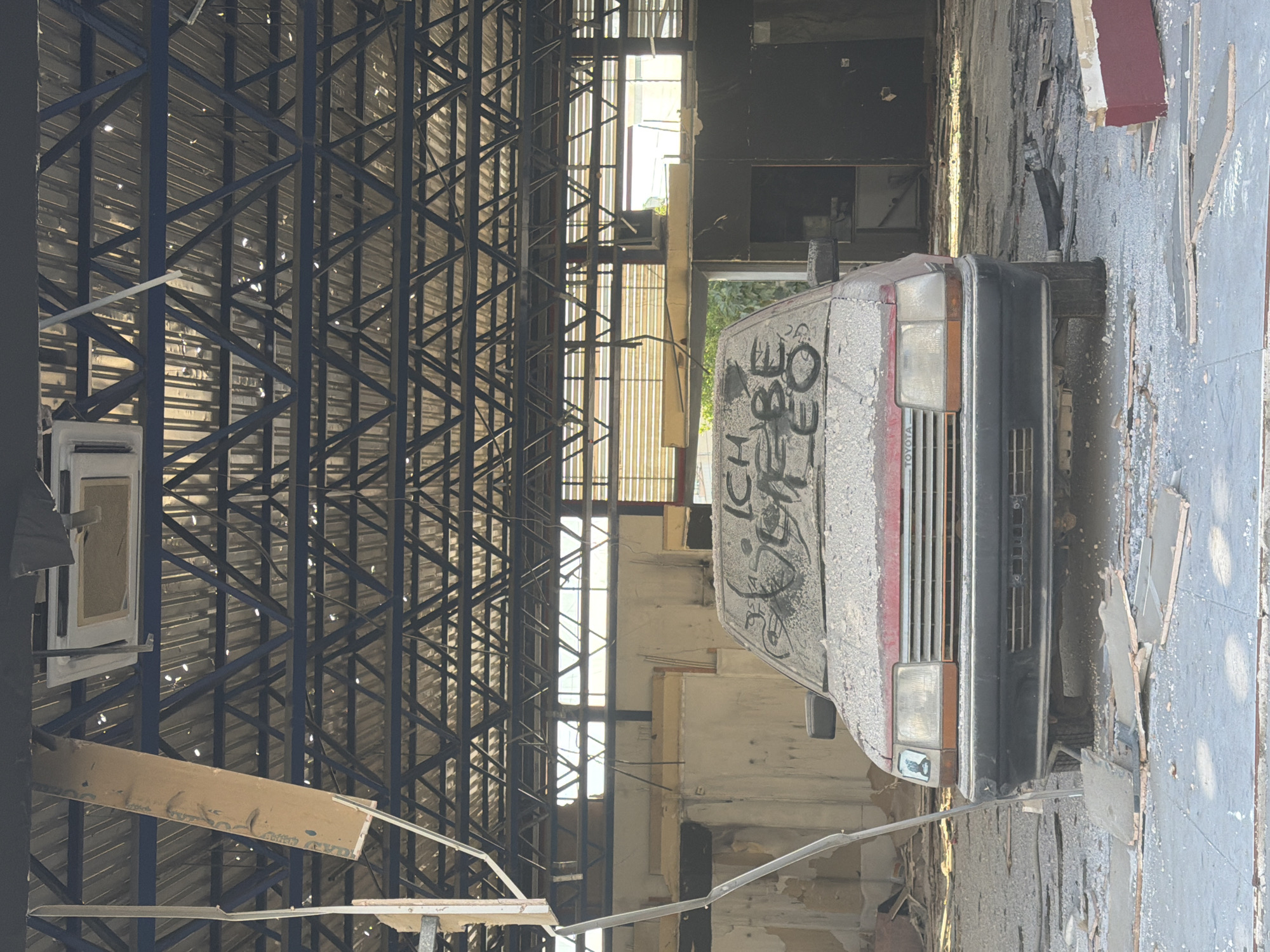 The image depicts an abandoned car in a derelict industrial building in Larnaca, Cyprus. The car, covered in dust and graffiti, appears to be a Toyota, judging by the partially visible logo on the front. The vehicle's front is facing the viewer, with the passenger side partially visible. The interior of the building is in severe disrepair, with debris scattered on the ground. The roof structure consists of blue metal beams and supports. Several windows allow sunlight to filter into the building. The overall environment suggests significant neglect and decay.