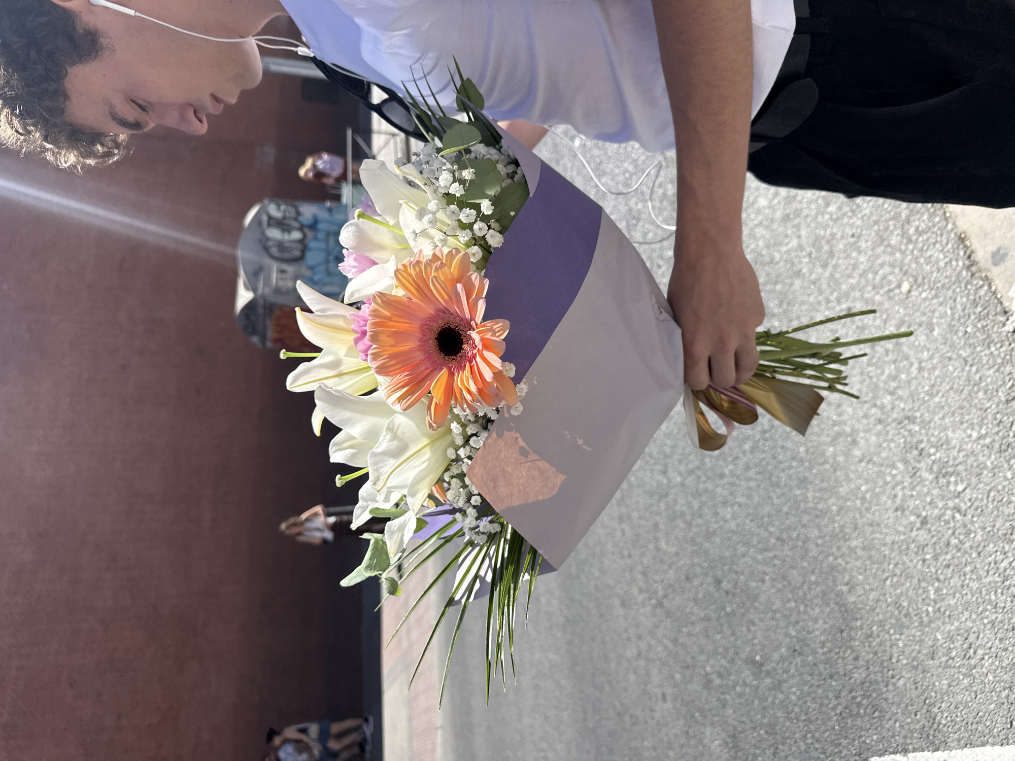 The image shows a young man holding a bouquet of flowers outdoors in an urban environment. The man, with dark curly hair and fair skin, is positioned on the left side of the frame, wearing a white t-shirt, black pants, and white wired earbuds. His head is slightly tilted, and his gaze is directed downwards or straight ahead.
He holds a vibrant bouquet in his right hand. The bouquet features several white lilies, a prominent orange gerbera daisy with a dark center, smaller delicate pink flowers, white baby's breath, and various green foliage. It is wrapped in a two-tone paper—light purple and white—and tied with a gold ribbon at the base.
The background on the left consists of a blurred reddish-brown building or wall. Further back, indistinct figures of other people and a blurry structure, possibly adorned with graffiti, can be seen, indicating a busy city setting. The ground is a light gray, textured pavement. The scene is brightly lit by natural sunlight, suggesting a clear, sunny day, potentially in Madrid, Spain, as per the location context. No specific event is depicted other than the man carrying the flowers. There is no visible text in the image, and the man's expression appears neutral.