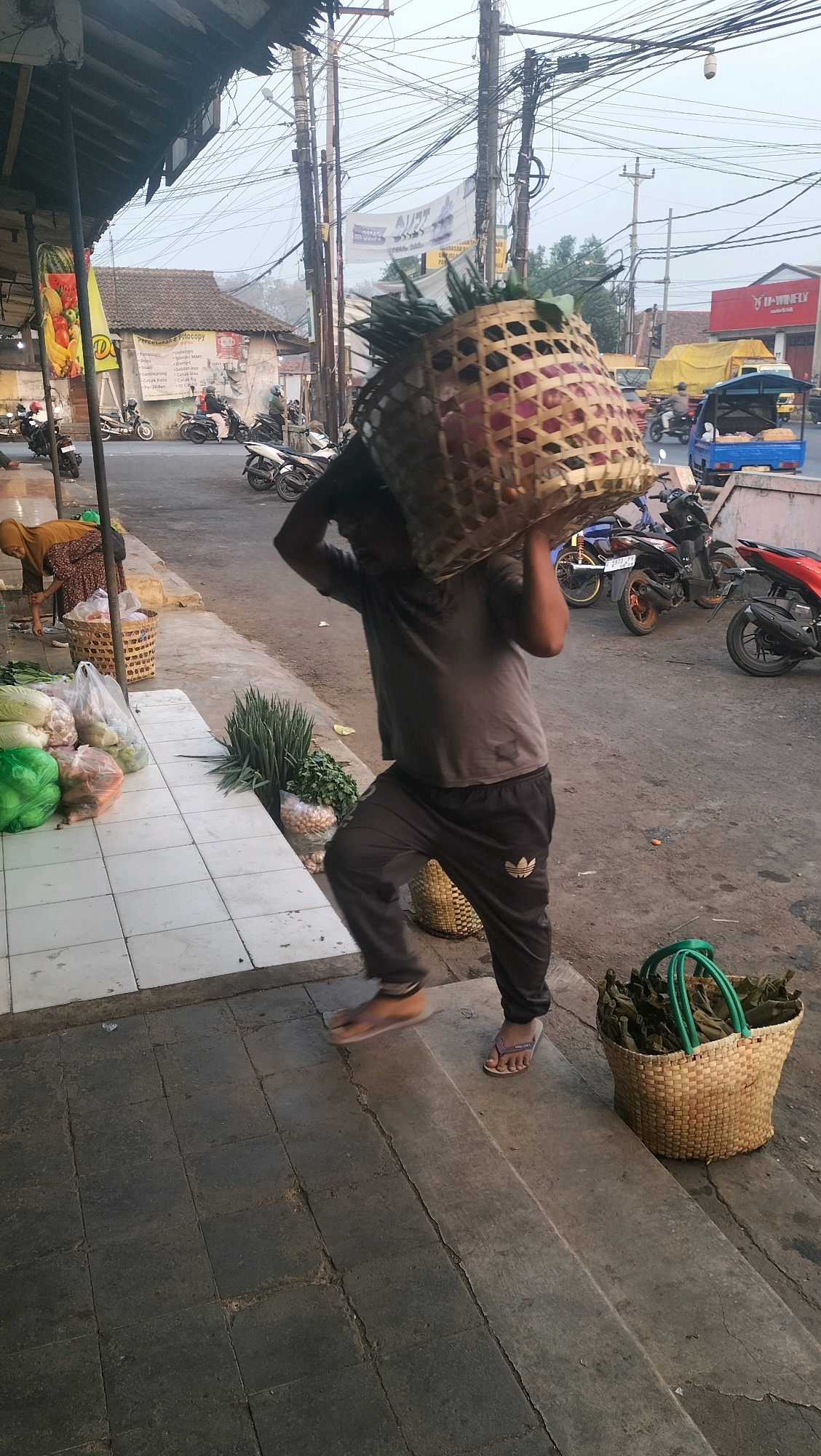 A man walks on a sidewalk carrying a large woven basket of produce on his head. The basket contains red onions and other vegetables. He is wearing a gray t-shirt and dark pants. The man is in a city setting with buildings, power lines, and vehicles visible. A woman is seen in the background near a stall with vegetables on display. In the foreground, there is a basket filled with dried leaves. The scene is likely in Subah, Indonesia.