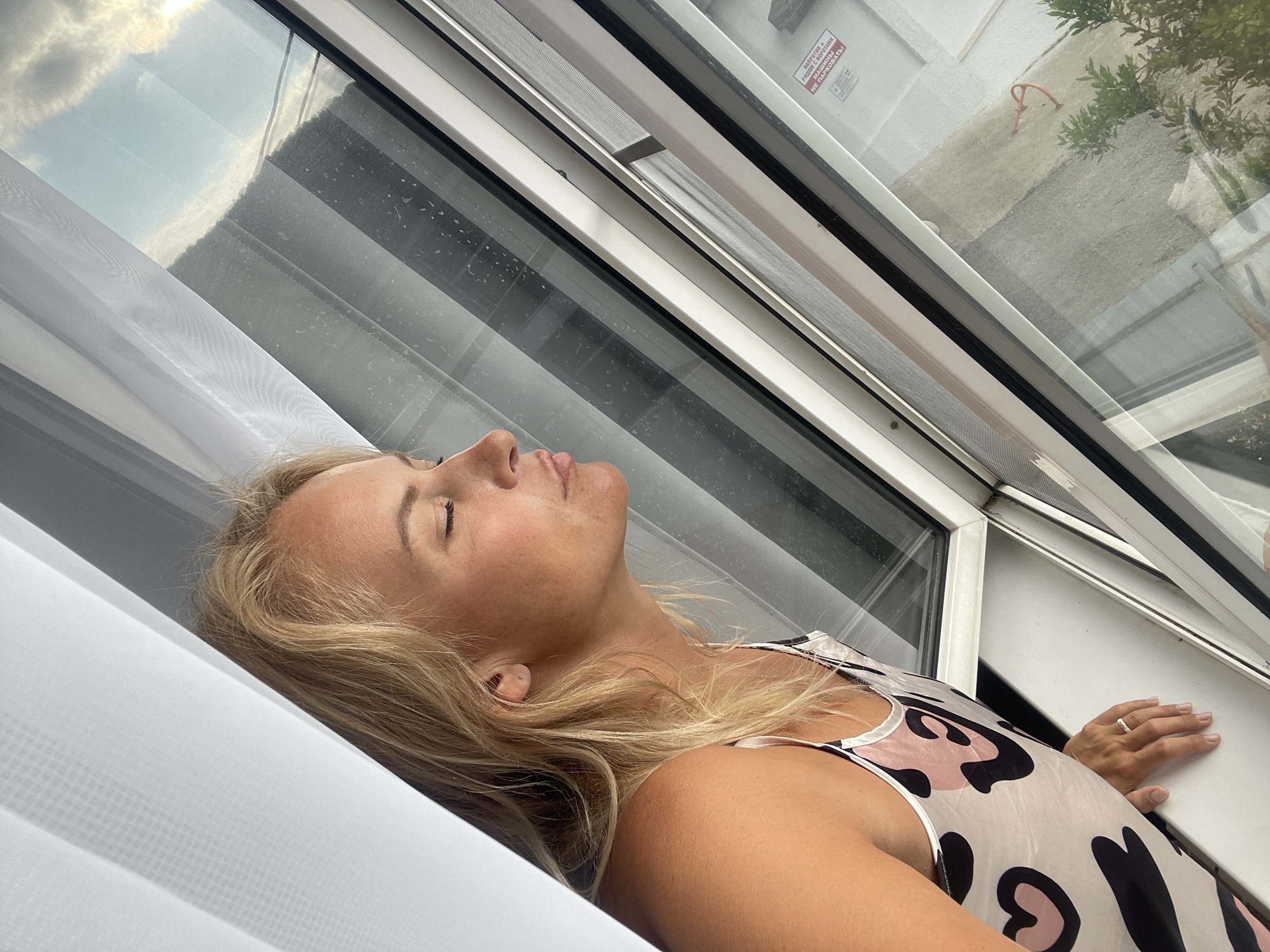 A blonde woman with her eyes closed and lips pursed is looking out of a skylight window. She is wearing a white tank top with a black and pink heart pattern. Her right hand is resting on the window sill, and she is wearing a ring on her finger. The window is open, and there are white curtains on the left side of the frame. The view outside the window shows a cloudy sky, some dark horizontal lines that could be blinds or part of the building's exterior, and some greenery. There is a warning sticker on the window frame. The scene appears to be indoors, likely in a home or hotel room, in the city of Baranovka, Russia.