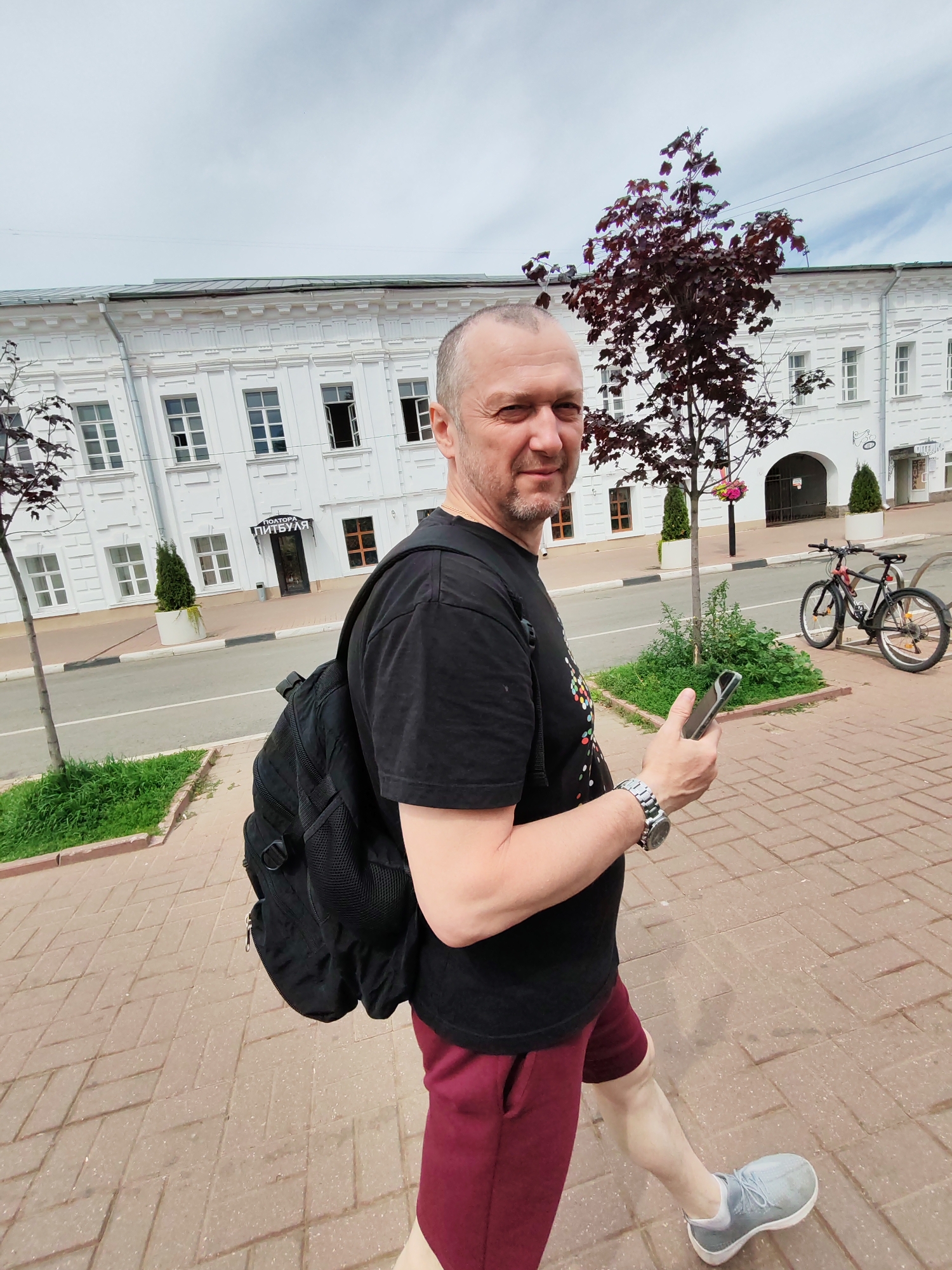 A middle-aged man with short, graying hair and a goatee walks on a brick-paved sidewalk in front of a white, two-story building. He is wearing a black t-shirt with a colorful, abstract design, maroon shorts, and gray athletic shoes. A black backpack is slung over his left shoulder, and he holds a silver smartphone in his right hand. The building appears to be a shop with an arched entrance and several windows. A sign above the entrance reads "ПИТБУЛЯ" (Pitbulya). Across the street, a bicycle is parked next to a bench. Trees with dark red leaves are planted along the sidewalk. The sky is cloudy. The man is looking at the camera with a slight smile. The setting is likely in a city, given the architecture and the presence of a street and parked bicycle. The sign "ПИТБУЛЯ" is a Russian word, suggesting the location is in Russia. The presence of trees and the architectural style may indicate a historic district. Yaroslavl, Russia, is a city known for its historic white stone buildings and well-preserved architecture, making it a plausible location for this scene.