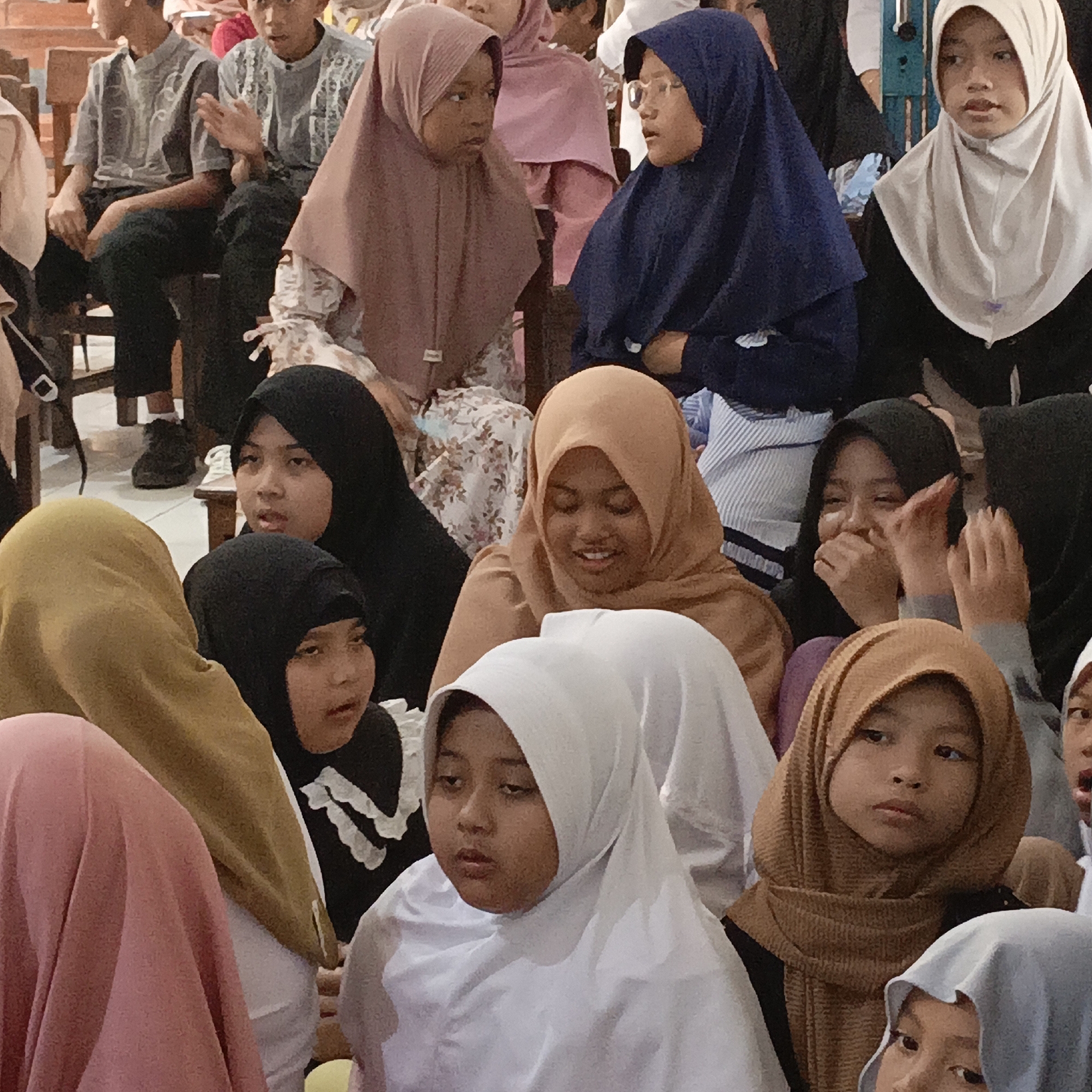 The image depicts a gathering of children, predominantly young girls, all wearing hijabs. They are seated in what appears to be an indoor setting, possibly a community hall or a classroom, indicated by the presence of wooden chairs and a tiled floor. The lighting suggests an interior environment, possibly with overhead illumination. The children are in various poses, with some looking forward, others in conversation, and some with their faces partially obscured. The overall atmosphere seems to be one of community or a religious gathering. While the location is stated to be Salatiga, Indonesia, there are no specific visual cues within the image to confirm this.