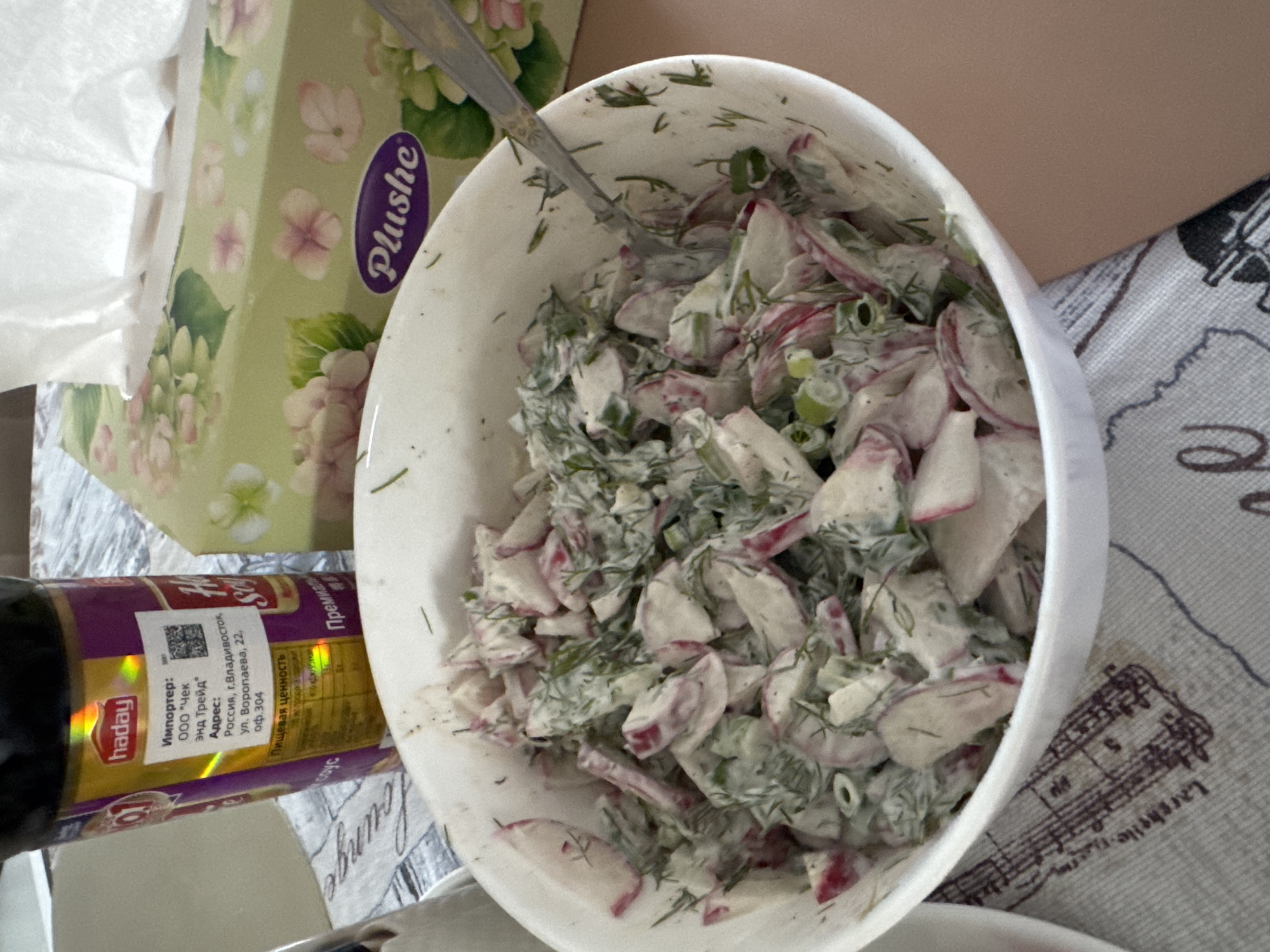 A close-up shot shows a bowl of radish salad on a patterned tablecloth. To the left, a bottle with a purple and yellow label that says "haday" is partially visible. To the right, a box of "Plushe" tissues with a floral design sits on the table. The salad appears to be made with sliced radishes, chopped dill, and a creamy dressing. A spoon is partially submerged in the salad. The background is blurred, but appears to be an indoor setting. No people are visible in the image. The location of Anapskaya, Russia, is not determinable from the visual information provided.