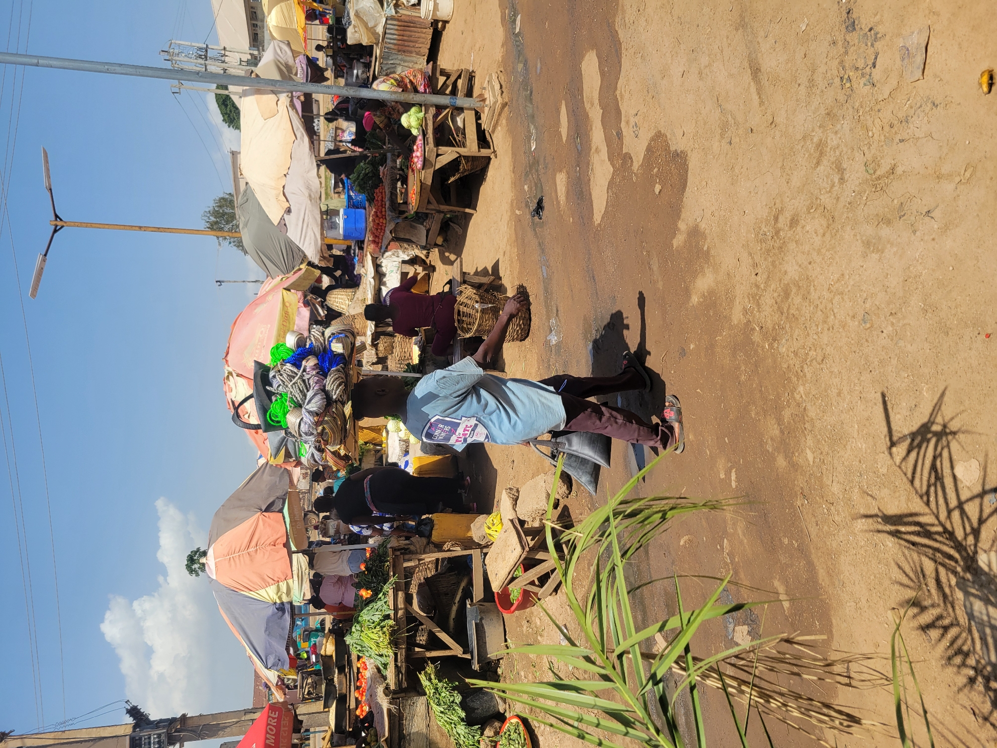 The image captures a bustling marketplace in Rafin Par, Nigeria. Stalls line the dirt ground, shaded by colorful umbrellas and filled with produce like tomatoes, greens, and other goods. People are browsing and interacting with vendors. A boy is walking through the marketplace carrying a container. The environment is sunny. A utility pole rises in the background and palm-like plants grow near the foreground.