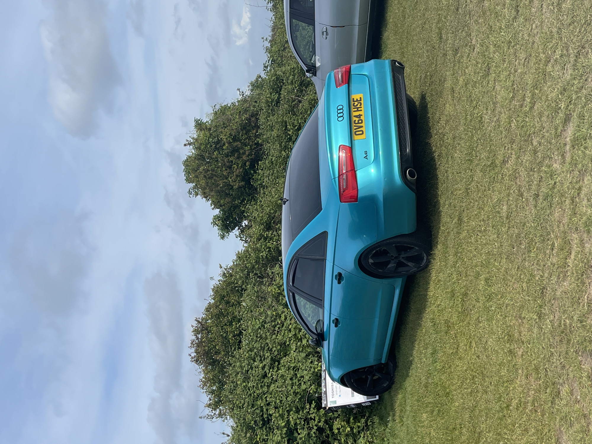 The image shows a turquoise Audi A6 parked on a grassy field with green foliage and trees in the background, under a partly cloudy sky. The car's license plate reads "OV64 HSE". Another vehicle, partially visible, is parked nearby. The location is possibly in Sevenoaks, United Kingdom, though no definitive landmarks are visible. There are no people visible in the image.