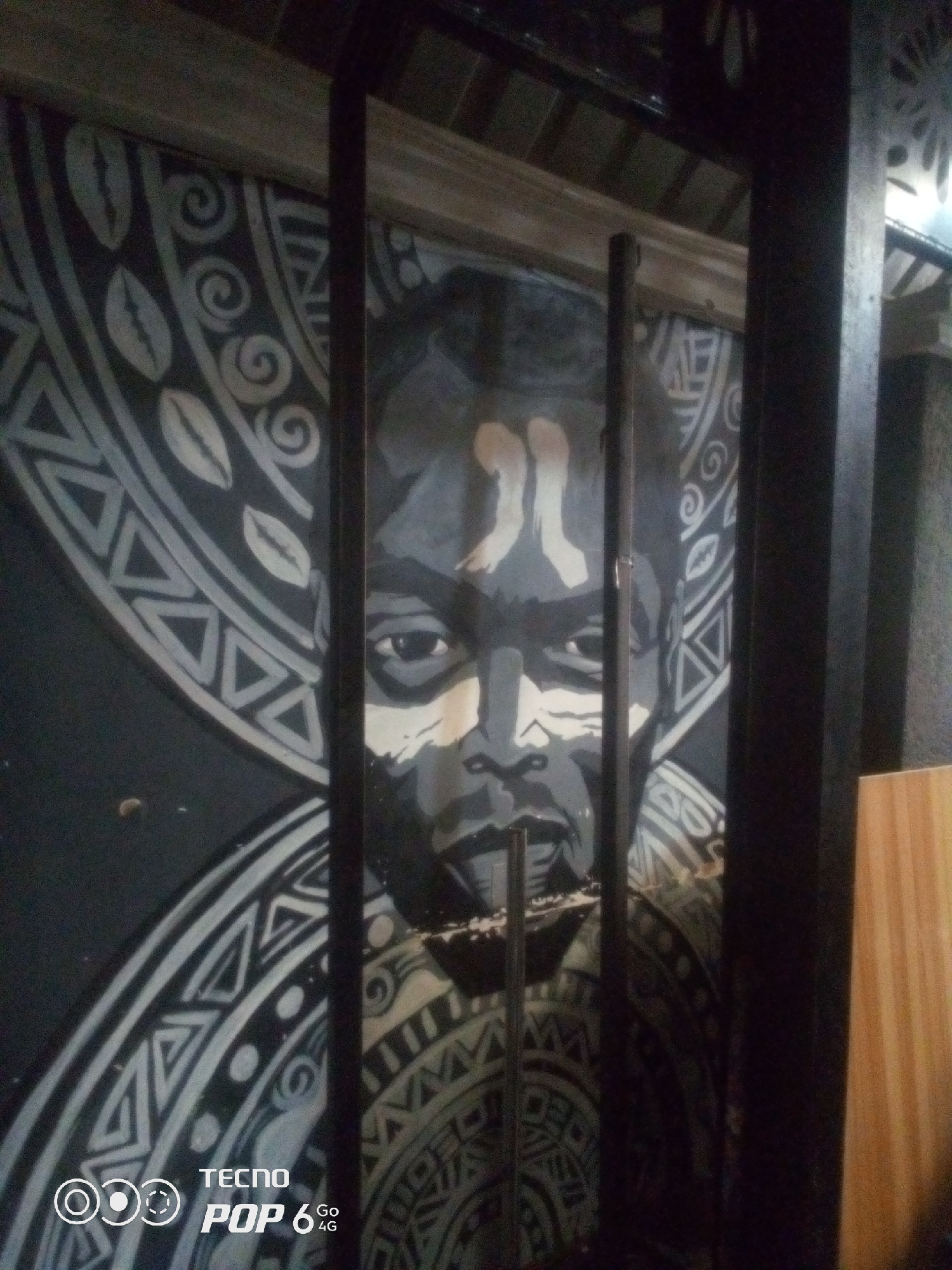 The image depicts a mural painted on a dark wall, possibly in an indoor establishment in Howayi, Nigeria. The mural features a stylized portrait of a person with dark skin, rendered in shades of black, white, and grey. The person has prominent eyes and facial features, with two light-colored markings on their forehead. The mural is framed by dark, vertical bars, which partially obscure the view of the artwork.
Surrounding the central portrait are intricate, circular patterns with geometric shapes and leaf-like motifs, also in black and white. These patterns create a tribal or culturally significant aesthetic. The background of the mural is dark, contrasting with the lighter elements of the design.
To the right of the mural, a portion of a light-colored wooden surface is visible, suggesting it might be a wall or furniture. In the lower left corner of the image, a Tecno Pop 6 4G logo is overlaid, indicating the device used to capture the photograph. The lighting in the scene appears to be artificial, casting some shadows and highlighting certain areas of the mural and its surroundings. The overall impression is that of a decorated interior space.
