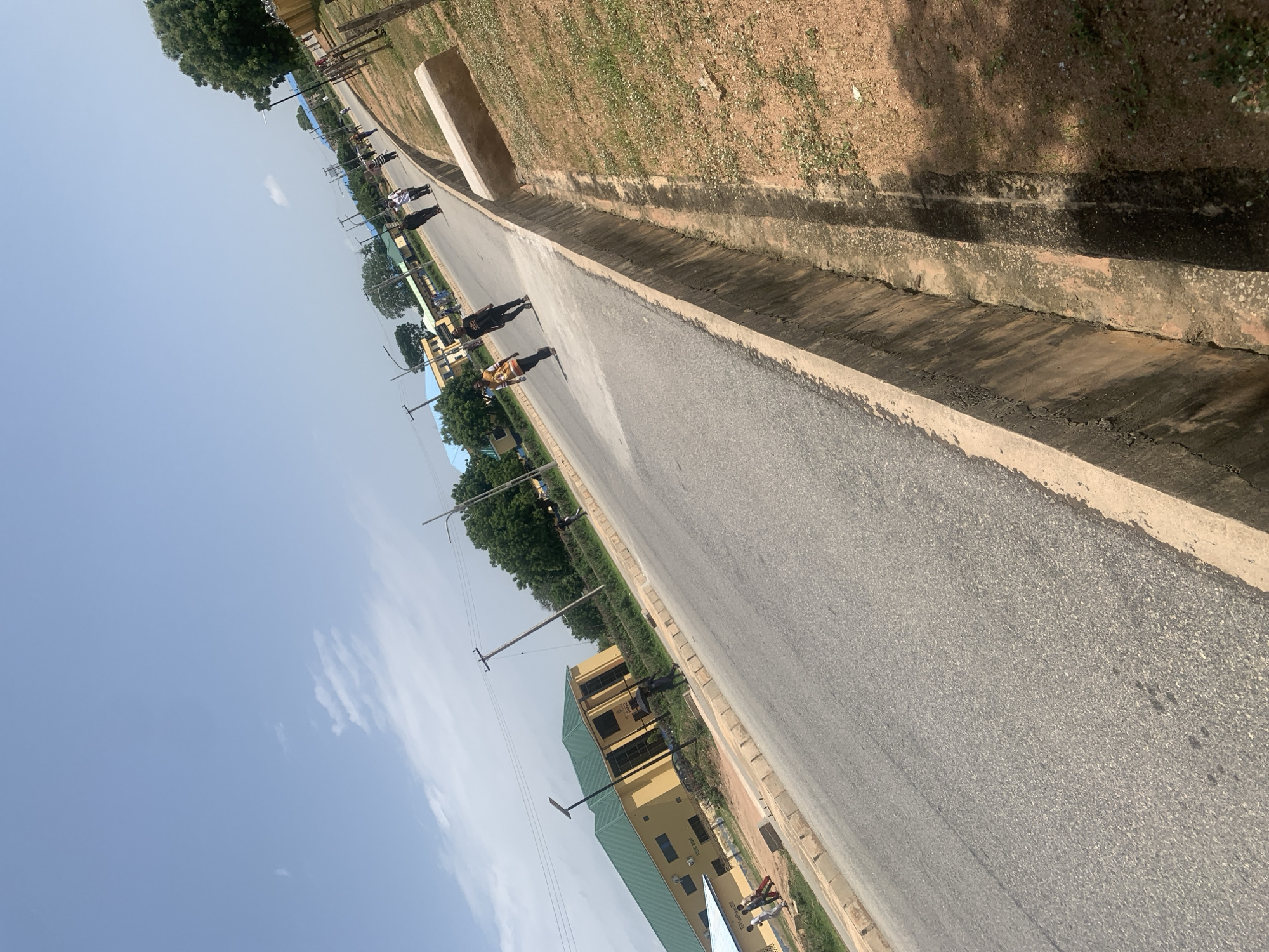 The image captures a street scene in Jalingo, Nigeria, under a bright, partly cloudy sky. A paved road dominates the foreground, with a concrete drainage ditch running along its right side. Several individuals are walking on the road, some appearing to be students or pedestrians. A yellow building with a green roof is visible on the left side of the road, likely a public or educational institution, with some people near its entrance. Trees and utility poles line the street, indicating an urban or semi-urban environment. The overall impression is that of a typical day in a Nigerian city, with everyday activities taking place.