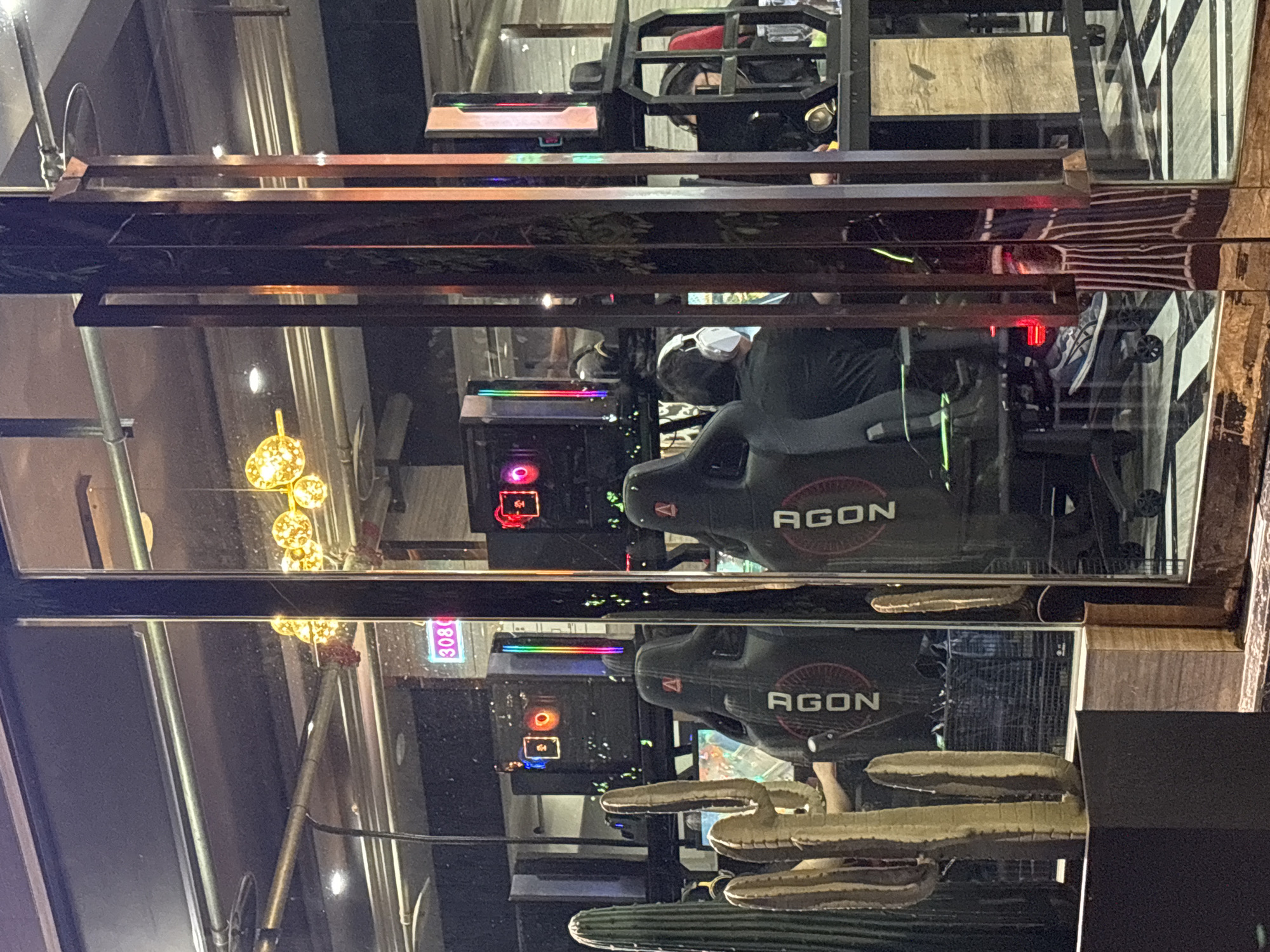 The image shows a commercial space with gaming setups, likely an internet cafe or eSports arena. The location is the Nan'an District in China. Several PCs are visible behind a glass or plexiglass barrier, illuminated with RGB lighting. One of the PCs has the number "3080" displayed in a neon-like sign. A black gaming chair with the brand "AGON" on it is in front of the PC. A person with headphones is sitting in the chair. Below the PC setup is a cactus. Above the PCs, another gaming setup is present.