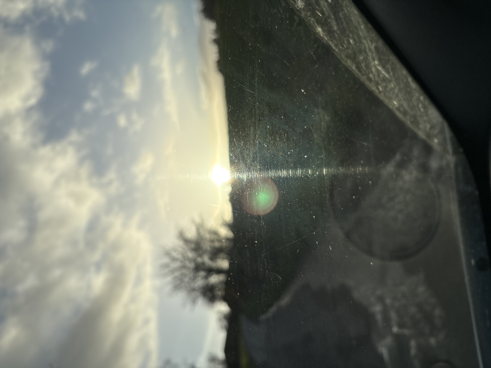 The image presents a rural landscape scene at low sun, viewed through a dirty and scratched vehicle windshield in Shelve, United Kingdom. The sun, positioned near the horizon slightly right of center, emits prominent lens flares, including a distinct green circular flare and vertical light streaks. A paved pathway or road curves from the lower-left foreground into the mid-ground, flanked by green grass and terrain. A cluster of leafless deciduous trees is silhouetted against a partially cloudy sky on the left horizon, with additional distant foliage or terrain visible on the right. The vehicle's interior, possibly a circular vent or dashboard element, is faintly reflected in the lower-center of the windshield. No people, animals, or specific interactions are present in the visible outdoor environment.