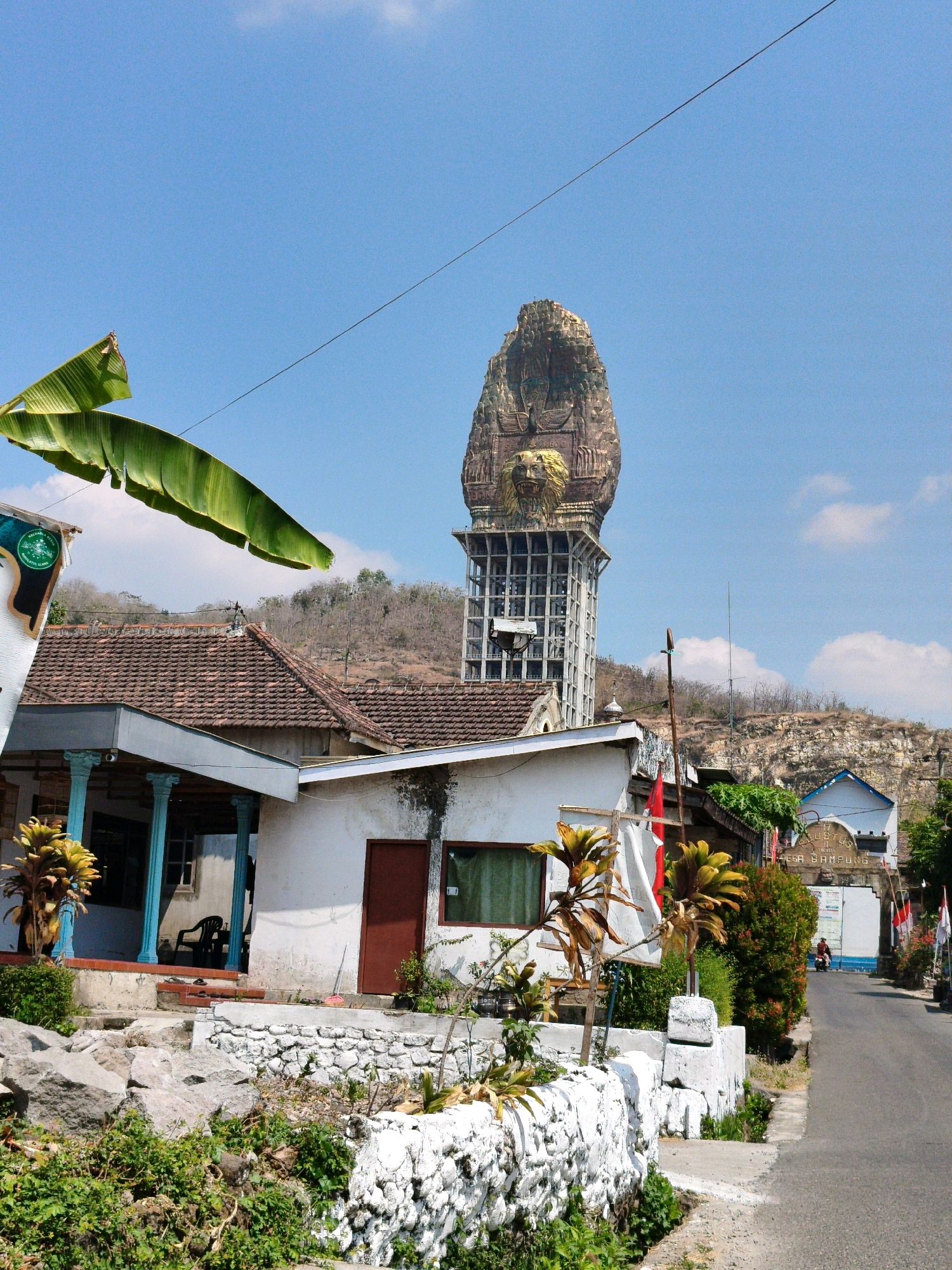 In the city of Sampung, Indonesia, a striking tower with a large, ornate sculpture atop it dominates the skyline. The sculpture depicts a stylized, serpent-like head with a golden lion's face at its base, emerging from a rocky structure. The tower itself is a multi-story concrete framework, suggesting ongoing construction or a unique architectural design. In the foreground, a white-painted stone wall borders a grassy area with scattered rocks. To the left, a white building with a tiled roof features light blue columns and a dark red door. Further down a paved road, another building with an archway displays text that appears to read "GUSTUS" and "DESA SAMPUNG." A single individual on a motorcycle travels away from the camera down the road. Lush green vegetation and some dry, spiky plants are visible throughout the scene, set against a clear blue sky with scattered white clouds. Banana leaves hang into the frame from the upper left corner.