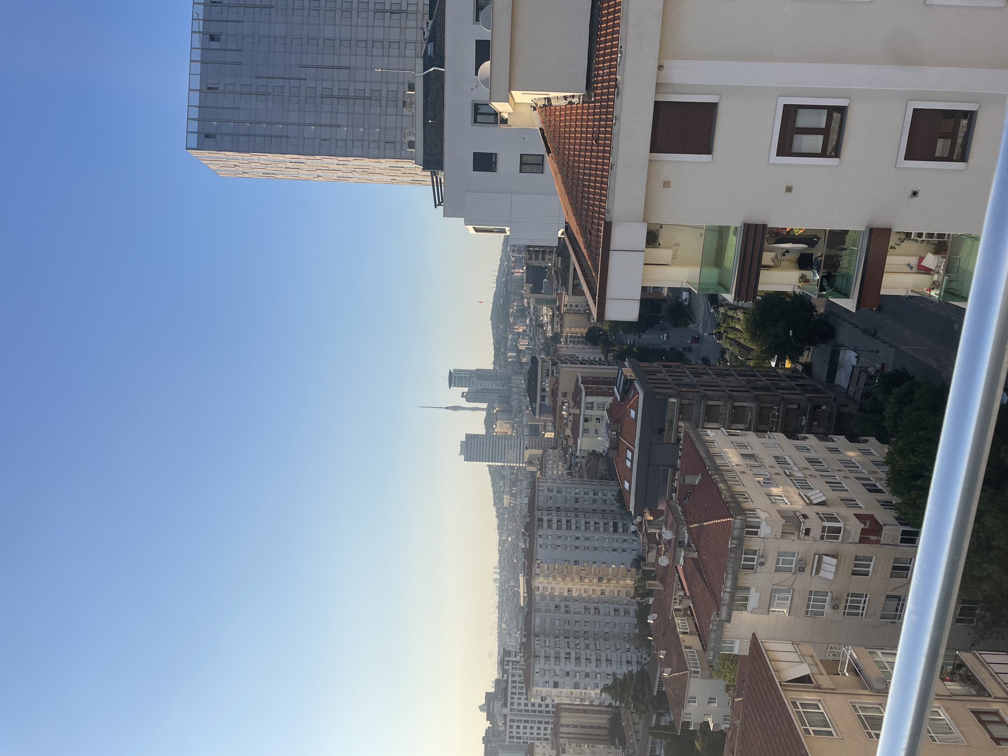 An aerial shot captures a cityscape in Kadıköy, Turkey, under a clear blue sky. The foreground is dominated by the edge of a modern building with a metallic railing, and to the right, a lighter-colored building with red-tiled roofs and several windows. Further back, a dense collection of apartment buildings with varying architectural styles and red-tiled roofs fills the mid-ground. In the distance, a cluster of taller skyscrapers and a prominent tower with an antenna rise above the horizon, against a hazy sky. The buildings appear to be residential and commercial, with visible balconies and windows. A street with some vehicular traffic is visible in the middle ground, winding between the buildings. The overall impression is of a sprawling urban environment bathed in natural light, suggesting it is either early morning or late afternoon.
