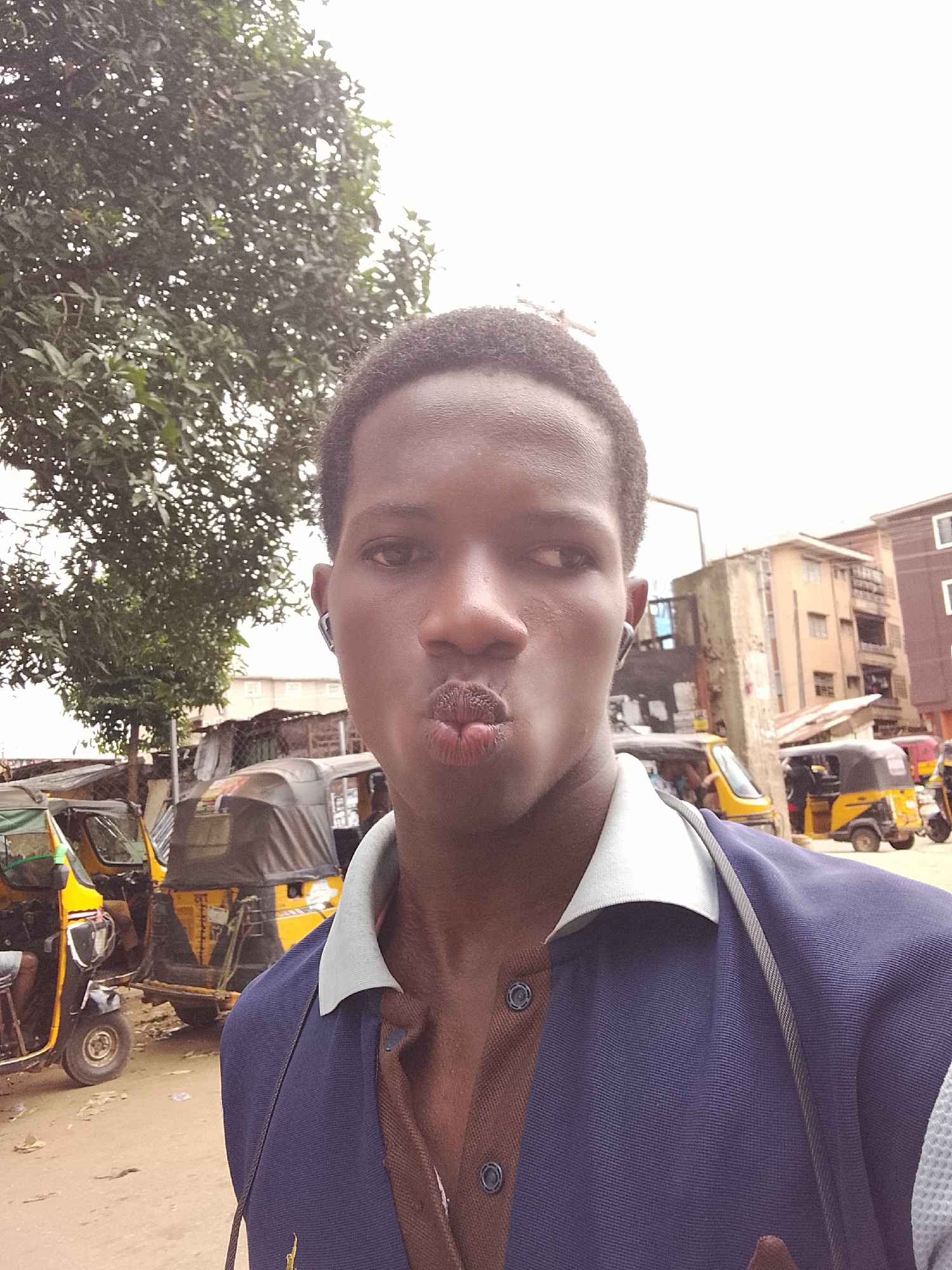 A young man, possibly a teenager, takes a selfie outdoors. He is in the foreground, his face filling the majority of the frame. He has dark skin, short dark hair, and is making a kissing face with his lips puckered. He is wearing a dark blue collared shirt over a brown shirt, with earbuds in his ears.
Behind him, the scene is busy and somewhat cluttered. Several yellow auto-rickshaws, commonly known as tuk-tuks, are parked on a dirt road. Some have covers over them, suggesting they are either for sale or being serviced. In the background, there are buildings, some of which appear to be multi-story with balconies. A large tree with green leaves is on the left side of the image. The sky is overcast, indicating it may be a cloudy day. The overall impression is of a street scene in a populated area, possibly a market or transportation hub in an African or Asian country. The time of day is not clearly discernible due to the overcast conditions, but it appears to be daytime. No visible text is present in the image.