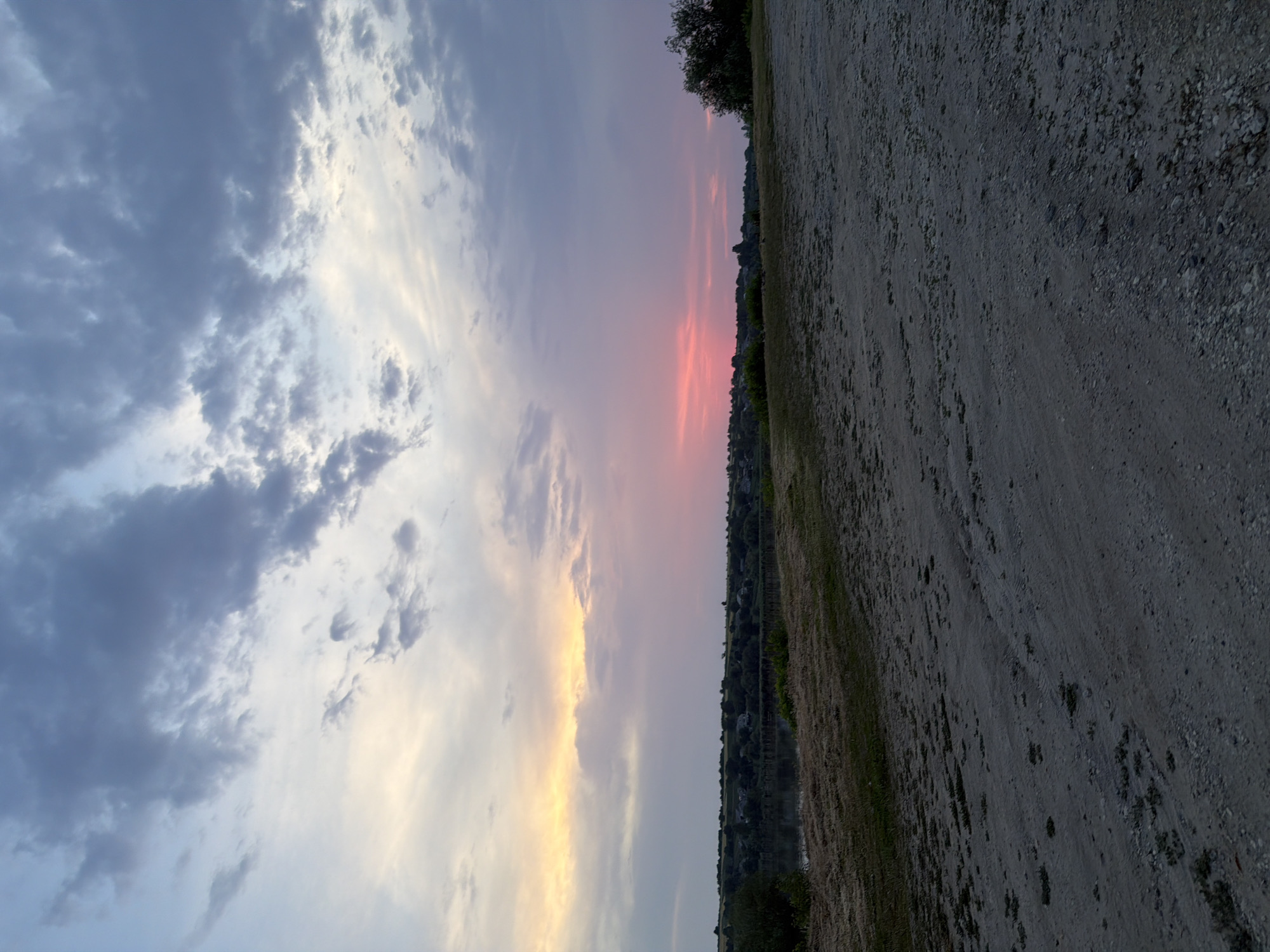 The image captures a dusky rural landscape under a dramatic sky. In the foreground, a wide expanse of dry, gravelly earth occupies the bottom half of the frame, showing tire tracks and sparse patches of dry grass. The middle ground features a gentle slope covered in dry, yellowed grass, leading down to a body of water, possibly a river or lake, with reflections visible. Beyond the water, a line of trees and scattered houses forms a distant horizon, suggesting a small settlement. The background is dominated by the sky, which transitions from soft, pale yellow and pink hues near the horizon to darker blues and grays with textured clouds higher up. The overall lighting indicates either sunrise or sunset, casting a serene and somewhat melancholic atmosphere. There are no people or animals visible in the frame.