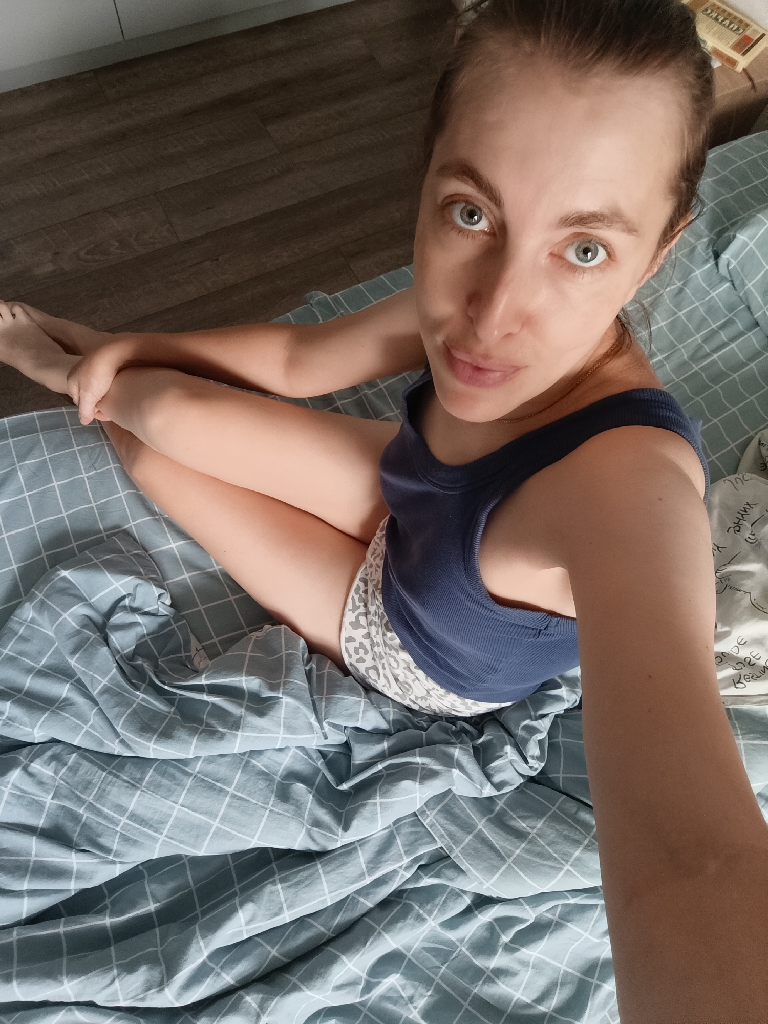 A woman is sitting on a bed, looking at the camera. She is wearing a blue tank top and patterned shorts. The bed is covered with a blue and white checkered duvet. The floor is made of dark wood planks. The room appears to be a bedroom, possibly in an apartment in the city of Chaikovsky, Russia. The woman's legs are crossed and her arms are resting on her knees. The lighting is natural, coming from an unseen window.
