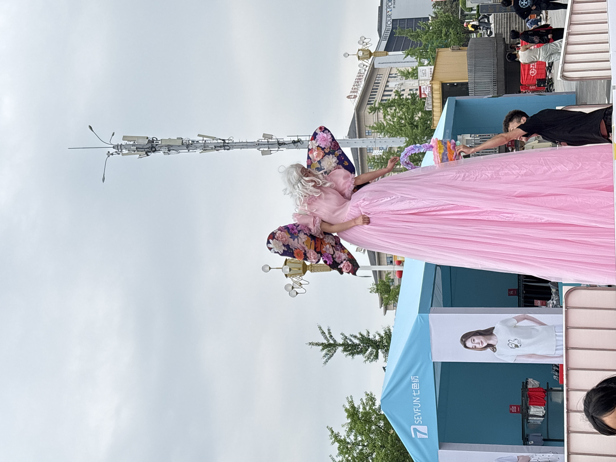 An outdoor event in Jiangjin, China, features a person in a pink gown with floral butterfly wings and white wig, holding out a colorful object to a person in a black shirt. A light blue tent with a SEVFON 七色纺 banner and a poster of a woman in a white shirt is nearby. The background includes buildings and street lights.