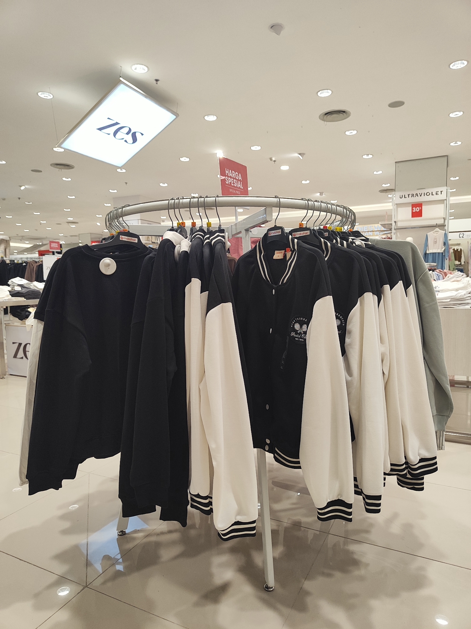 A clothing store display features a circular rack of jackets and sweatshirts. The items are predominantly black and white, with some in grey and light blue. Some garments have striped cuffs and collars. One jacket is black with white sleeves and features an embroidered logo with a tennis racket and ball. In the background, various other clothing racks and displays are visible, along with ceiling lights. A sign above the clothing reads "ZES." Another red sign with "HARGA SPESIAL" and "SPECIAL PRICE" is also present. A sign with "ULTRAVIOLET" and "30%" is visible in the background. The floor is tiled and reflective. This scene is located within a Matahari department store in Nangkaan, Indonesia.