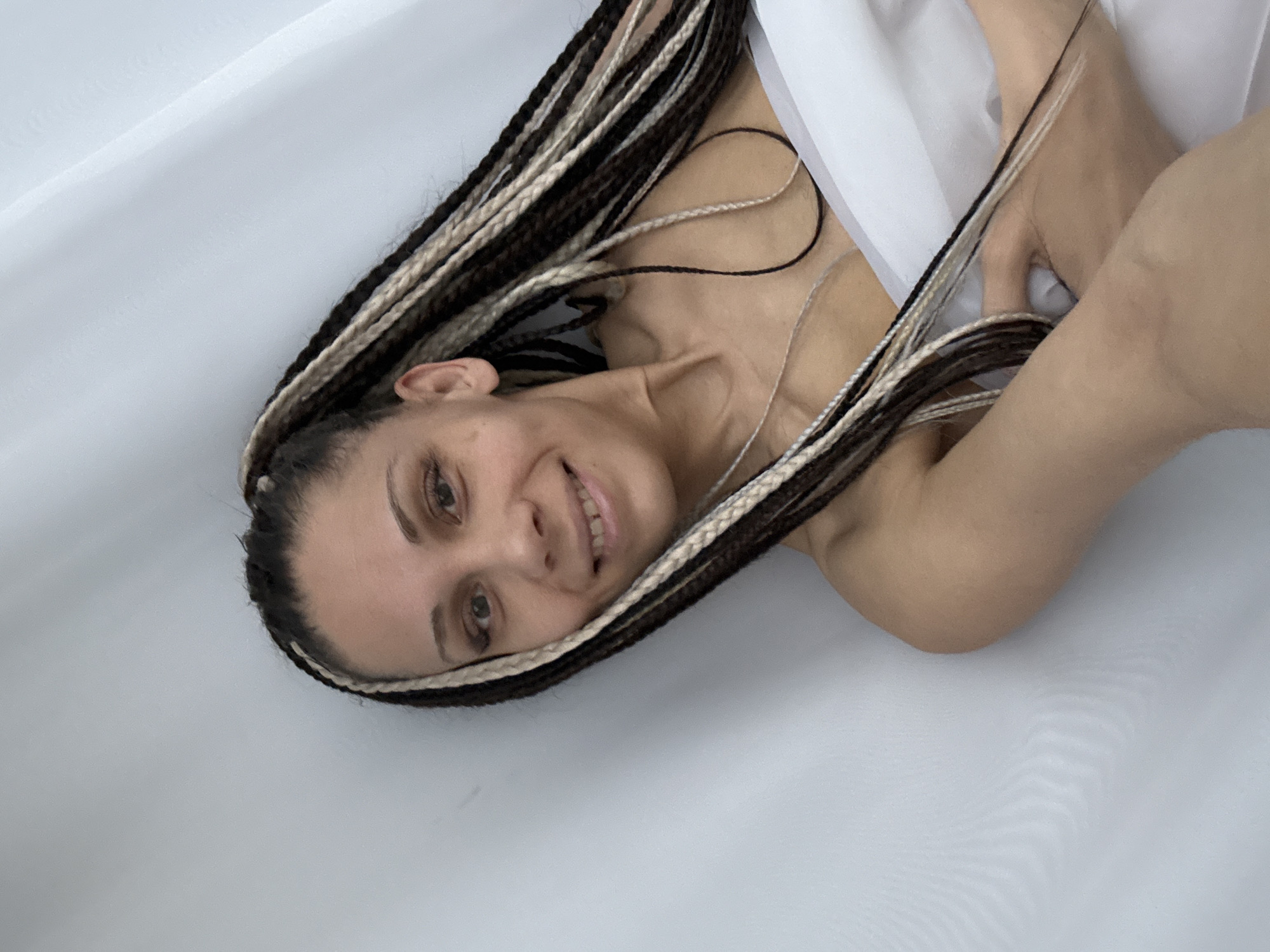 A smiling woman with multi-toned braids (dark brown, light brown, and white/blonde) lies on a white surface, looking directly at the camera. Her head is tilted to the right, and her long braids cascade around her head and shoulders. She has a wide, cheerful smile, revealing her teeth, and her eyes appear light brown or hazel. A small stud piercing is visible on her left nostril. Her bare shoulders and upper chest are visible, partially covered by a soft, white fabric that drapes around her. One arm is bent, with her hand near her chest. The background consists solely of plain white material, suggesting a studio setting, bed, or a minimalistic environment. The lighting is soft and even, highlighting her features. The overall mood conveyed is friendly and relaxed. No text is visible in the image. The scene is set in Rusanovo, Russia.