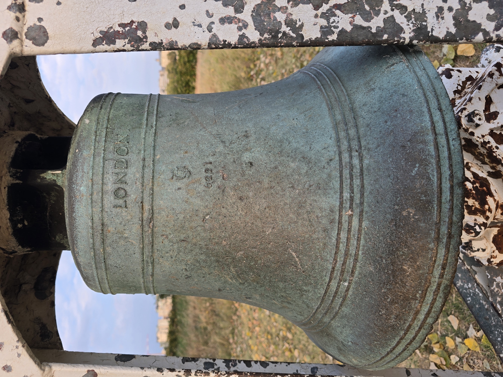 The image shows a close-up, eye-level, vertical shot of a weathered, verdigris-coated bronze bell. The bell is mounted within a white metal frame, which displays significant rust and chipping. The text "LONDON" is clearly visible in raised lettering near the top of the bell, followed by a crest and the number "6221." The bell exhibits horizontal banding near its base. The background is softly blurred, showing dry, yellowish grass, some fallen autumn leaves, and a distant line of buildings under a partly cloudy sky. There are no people or animals present, and no actions are depicted. The scene appears to be outdoors, possibly in a park or open area, and the architectural style of the distant buildings suggests an urban or semi-urban setting. The description does not contain any information about Antwerp, Belgium.