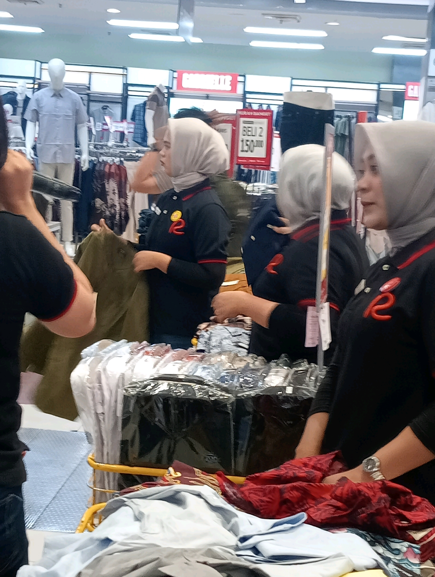 This is a medium shot, eye-level, indoor photograph taken in Jambi, Indonesia. The scene appears to be a retail store, possibly a department store or a supermarket, with clothing racks and displays visible in the background. The lighting is bright, consistent with overhead retail lighting.
In the foreground and midground, three women are the primary subjects. Two of the women are wearing dark polo shirts with red trim and a red logo on the chest, along with light-colored headscarves, indicating they are likely employees. One of these women, on the right, is in profile, looking towards the right edge of the frame. The other employee, in the center, is facing towards the left and appears to be interacting with a rack of folded clothes. She is holding a dark green garment. A third person, partially visible on the far left, is wearing a black t-shirt with red trim on the sleeves and is facing towards the right, seemingly interacting with the employee in the center.
The background shows various clothing items on display, including mannequins dressed in shirts and pants. There are also promotional signs, one of which is partially visible on the right and displays "BEI 2 150.000", suggesting a "Buy 2 for 150,000" promotion. Racks of clothing are also visible in the background, with items neatly hung or folded. The floor appears to be tiled. The overall impression is a busy retail environment during a sale or promotional event.