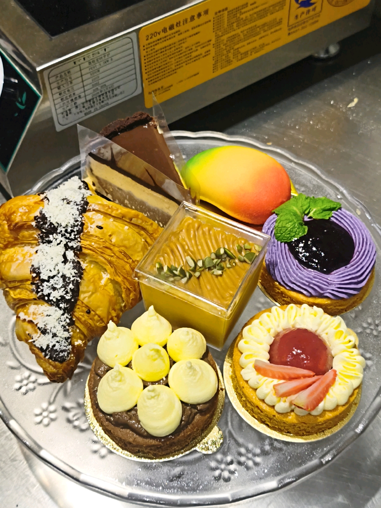 A close-up shot displays a clear glass platter filled with an assortment of delectable desserts, set against a stainless steel background. The platter is arranged with a chocolate cheesecake slice, a croissant generously dusted with shredded coconut and drizzled with dark chocolate, a rectangular cup of yellow custard topped with pumpkin seeds, a vibrant mango-shaped dessert with gradient colors of yellow, orange, and red, a purple tart with a swirl of cream and a dollop of blueberry jam crowned with mint, a chocolate tartlet adorned with eight dollops of yellow cream, and a round tartlet topped with yellow cream swirls, a red center, and sliced strawberries. The desserts are positioned on a table, implying an indoor setting. The overall scene is illuminated by soft lighting, highlighting the textures and colors of the pastries.