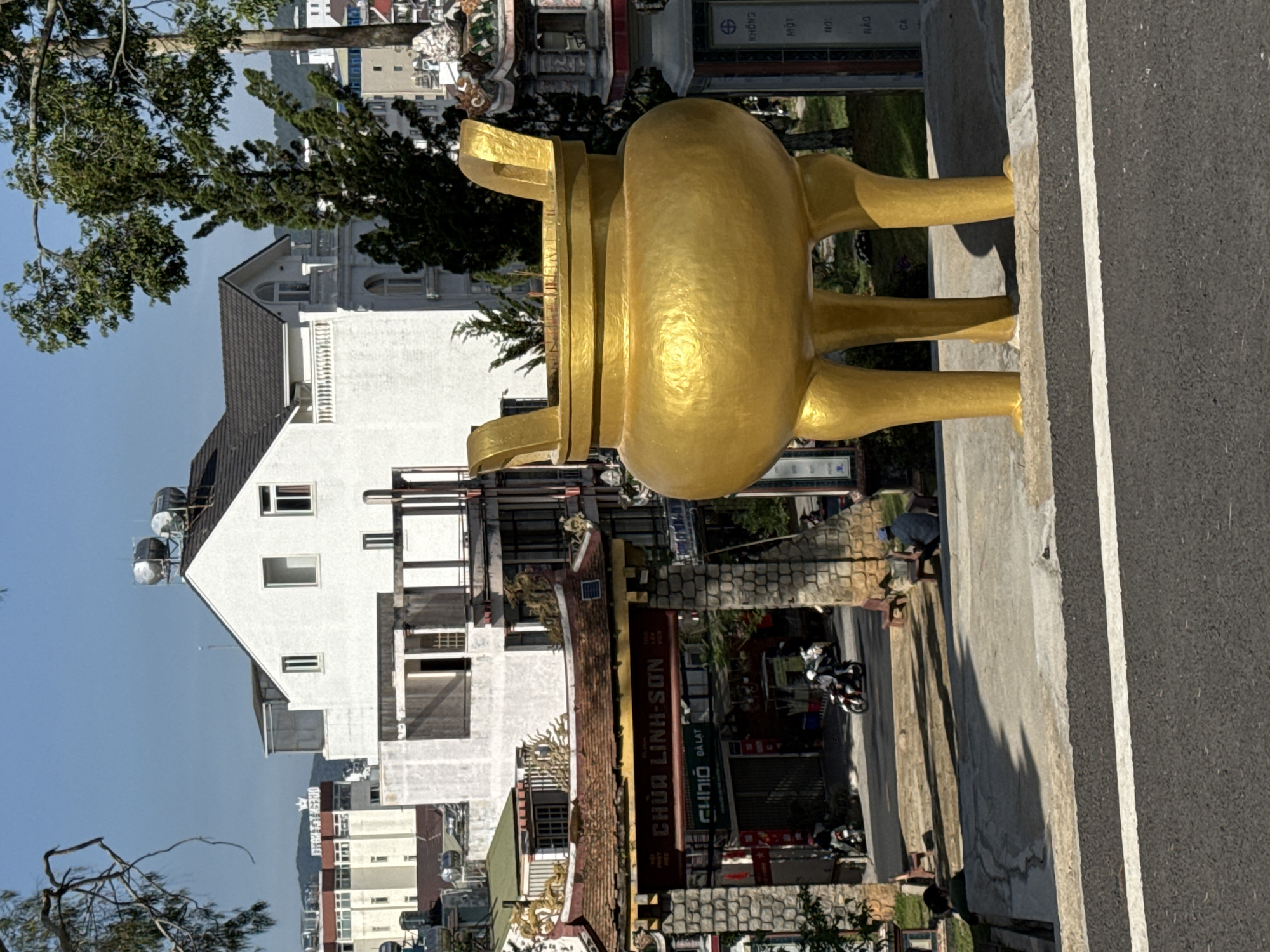 A vertical shot depicts a sunny street scene in Da Lat, Vietnam, featuring a large, gold-colored, metal incense burner in the foreground. The burner is situated on a gray concrete ledge adjacent to an asphalt road with a white dividing line.
Across the street, there's a row of buildings, including a white, multi-story house with solar water heaters on its roof. A sign that reads "CHÙA LINH-SƠN DA LAT" in Vietnamese is visible on a building. People and motorcycles are seen on the street.
The background shows more buildings and trees against a clear blue sky.