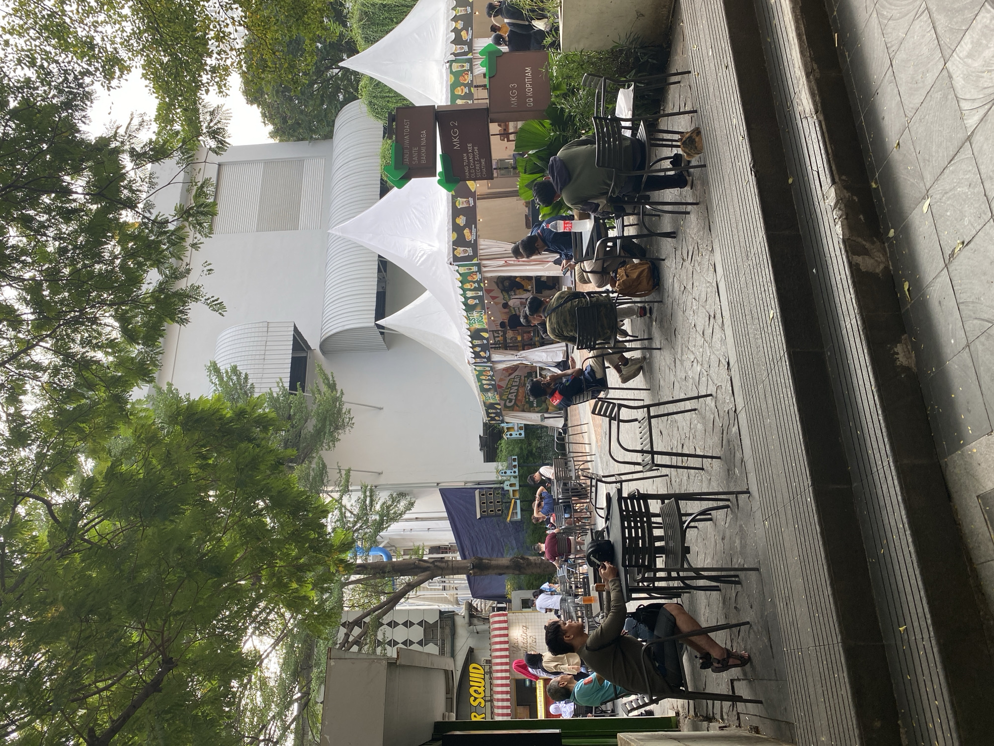 In a shaded outdoor seating area, people dine and socialize at metal tables and chairs. A white building with arched awnings stands in the background, while a large tree with lush green leaves provides shade. Several white tents are set up, likely for food stalls, with signage indicating different vendors. One signpost reads "MKG 2" and lists various food and drink options like "CHATIME". Another sign above it indicates "JANJI JIWA TOAST", "SANTE", and "BAKMI NAGA".
In the foreground, a man in a dark green shirt and shorts sits at a table, looking at his phone. Another man in a similar outfit is seated across from him. Further back, a group of individuals are engaged in conversation at a table, while others are seen milling about or seated in the tents. The ground is paved with interlocking tiles, and a set of concrete steps leads down to a lower level. The overall scene suggests a casual gathering or dining experience in an urban setting. This location is likely in North Jakarta, Indonesia, as indicated by the "MKG" signage, which often refers to Mall Kelapa Gading.