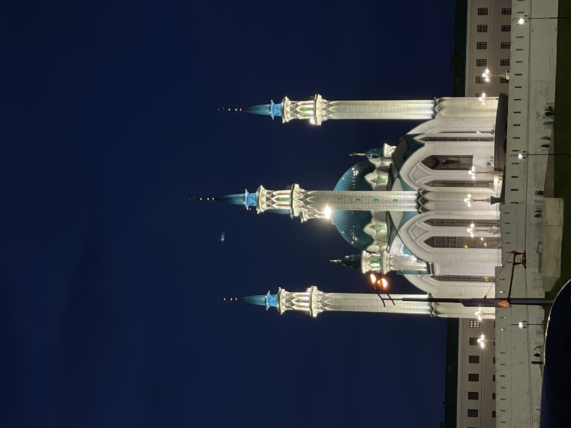 The image depicts the Kul Sharif Mosque in Kazan, Russia, at night. The mosque is illuminated, showcasing its white facade, turquoise domes, and four tall minarets topped with crescent moons. The sky is a deep, dark blue, suggesting nighttime. In the foreground, a white fortified wall encloses the mosque complex, with streetlights and a few figures visible along its parapet. Behind the mosque, a long building with multiple windows and a green roof is partially visible. The overall scene is one of architectural grandeur set against the dark night sky.