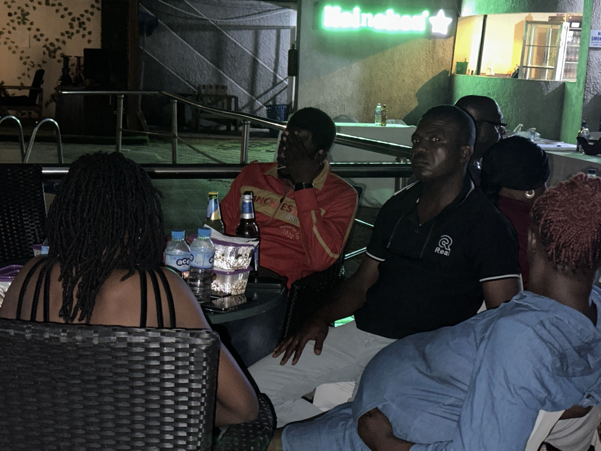The image depicts a group of people gathered around tables at an outdoor establishment, possibly a bar or restaurant, in Barden Yana, Nigeria. In the foreground, a woman with dark braided hair is seated with her back to the camera, facing a table with two bottles of water and some containers. To her right, a man in a red jacket is covering his face with his hands, suggesting distress or an attempt to hide. Next to him, a man in a black polo shirt with the "Real" logo is seated. Further back, other individuals are visible, including a woman with dark hair and a man with glasses. The environment appears to be dimly lit, with neon signage in the background indicating "Heineken" and a star. The overall scene suggests a social gathering in a public space.