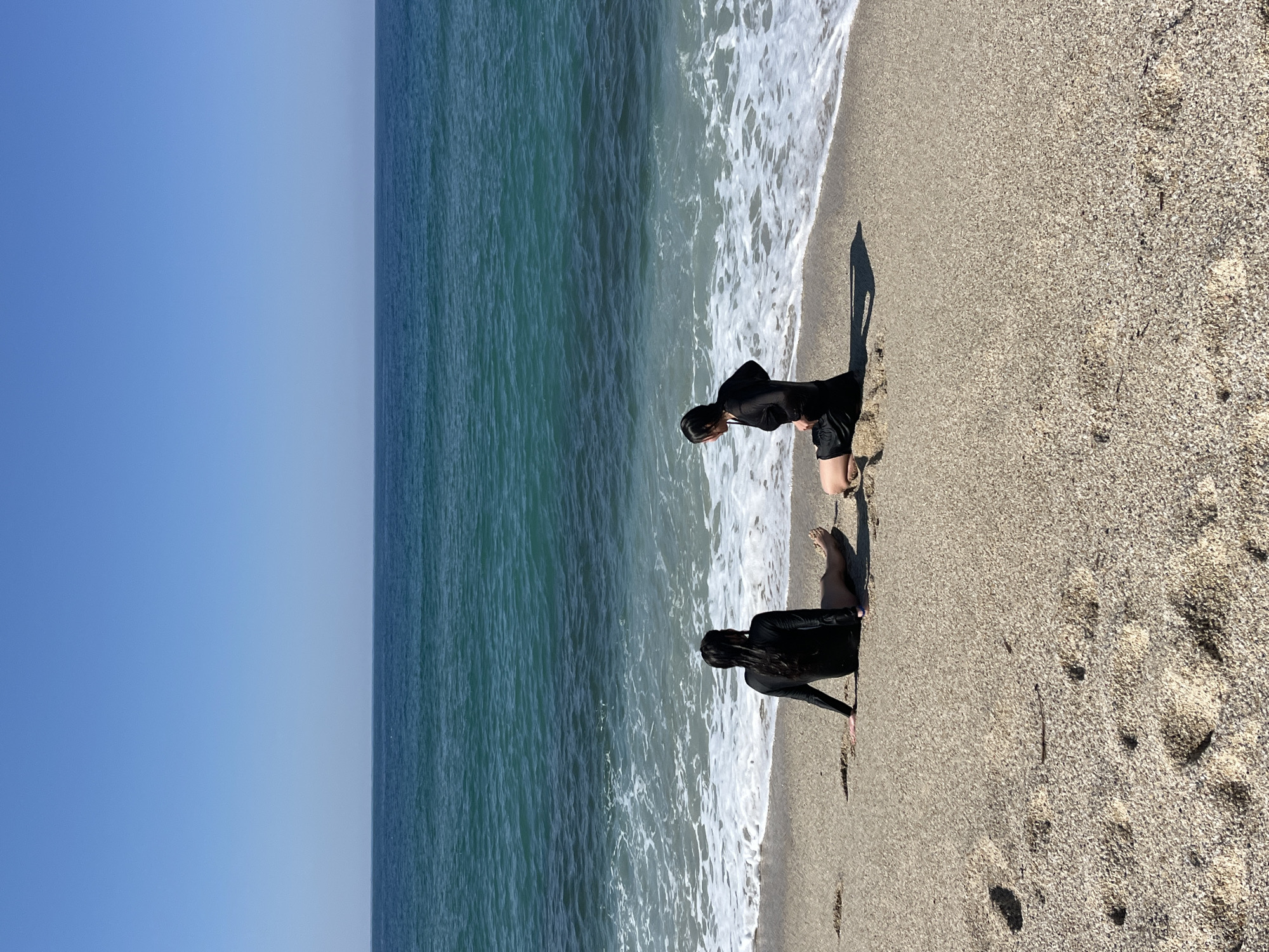 Two people are kneeling at the edge of the sea on a sandy beach. They are wearing dark clothing and facing the water. The location appears to be a beach in Alanya, Turkey. The sky is clear and blue, and the sea is a mix of blue and green with white waves. The beach extends to the right of the frame and is covered in sand and small pebbles.