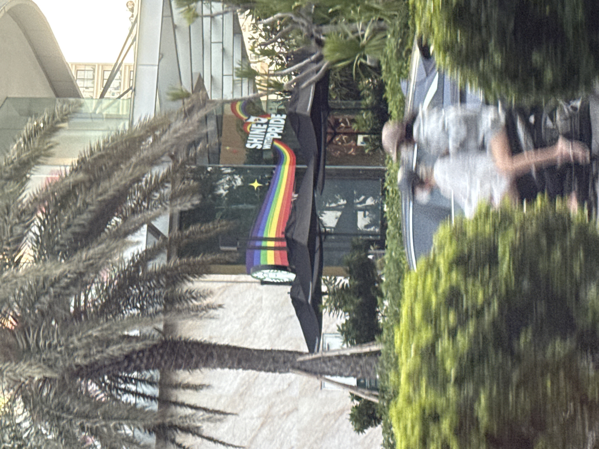 A medium shot captures a street scene in Nha Trang Ward, Vietnam, during daylight. A prominent palm tree on the left partially obscures the facade of a modern building with a glass front. A Starbucks store, identifiable by its black umbrellas and signage, is visible. The Starbucks logo is adorned with a rainbow-colored banner, and the text "SHINE WITH PRIDE" is displayed. In the foreground, a motorcycle carrying two people is captured in motion, their figures blurred by the movement. The motorcycle is passing through a section of lush green bushes. The background shows the upper levels of buildings, including windows.