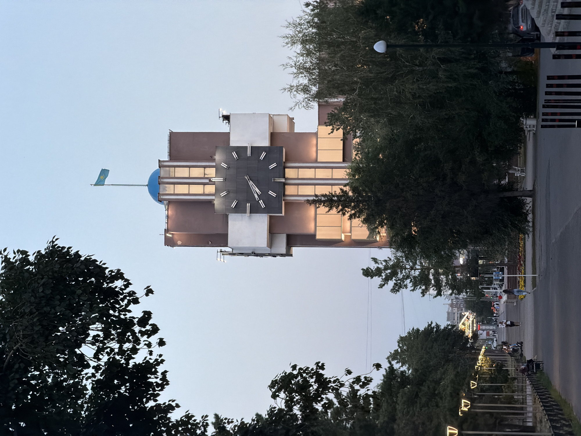 The image depicts a view of the clock tower in Pavlodar, Kazakhstan. The tower, with its prominent clock face and a blue dome topped by the Kazakh flag, stands against a pale sky. The building's facade is a muted brown with white architectural elements and illuminated windows. Trees frame the tower on either side, and a pathway with streetlights runs in the foreground, leading toward a street lined with more trees and some distant buildings. Several people are visible walking along the pathway, and a few are seated on benches. The overall scene suggests late afternoon or early evening, with the lighting indicating artificial illumination beginning to activate.
