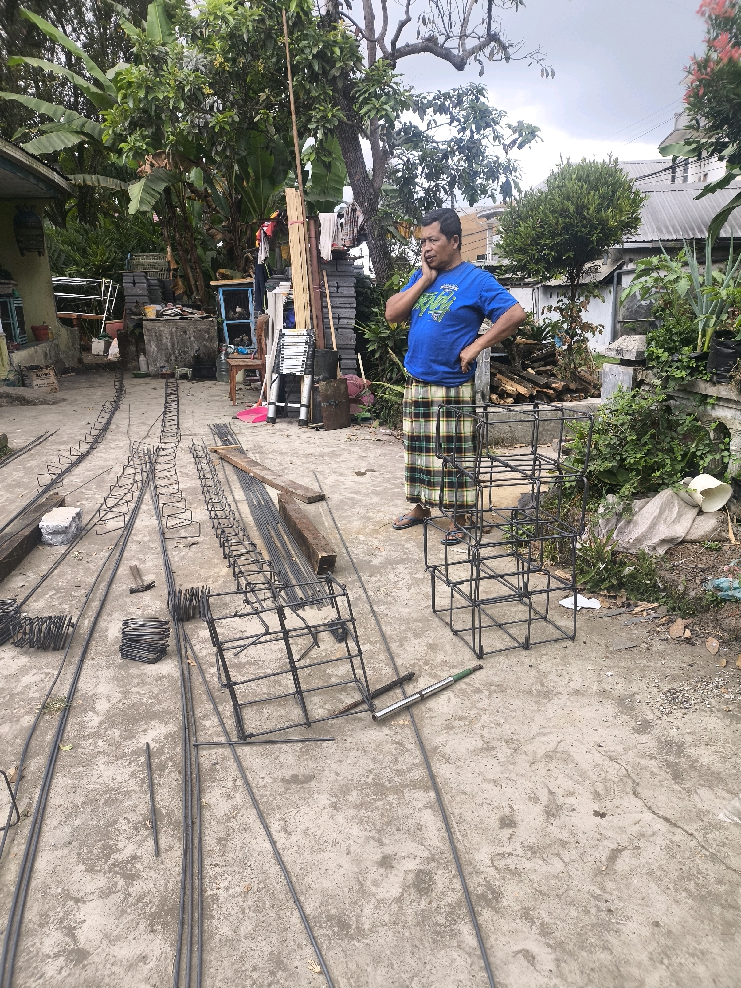 A man stands on a concrete surface in an outdoor area, possibly a yard or construction site, in Kandangtepus, Indonesia. He is wearing a blue t-shirt with a graphic design and a checkered sarong. The man is holding a mobile phone to his right ear with his right hand and has his left hand on his hip. He appears to be in conversation. The ground is covered with various construction materials, predominantly steel reinforcement bars (rebar) of different sizes and configurations. Several rebar cages, pre-bent into rectangular and cube shapes, are present. Bundles of rebar are also scattered around, along with some wooden planks and a concrete block. In the background, lush green foliage from trees and plants suggests a tropical or subtropical environment. A portion of a building with a grey roof is visible to the right. The overall scene depicts a man at a site where construction or groundwork is taking place.