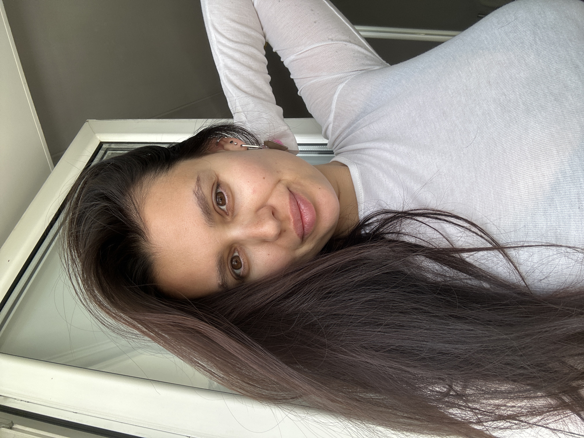 A woman with long, dark brown hair is lying down, close to a window frame, and wearing a white long-sleeved shirt. Her face is angled towards the camera. The location details are not discernible from the image.