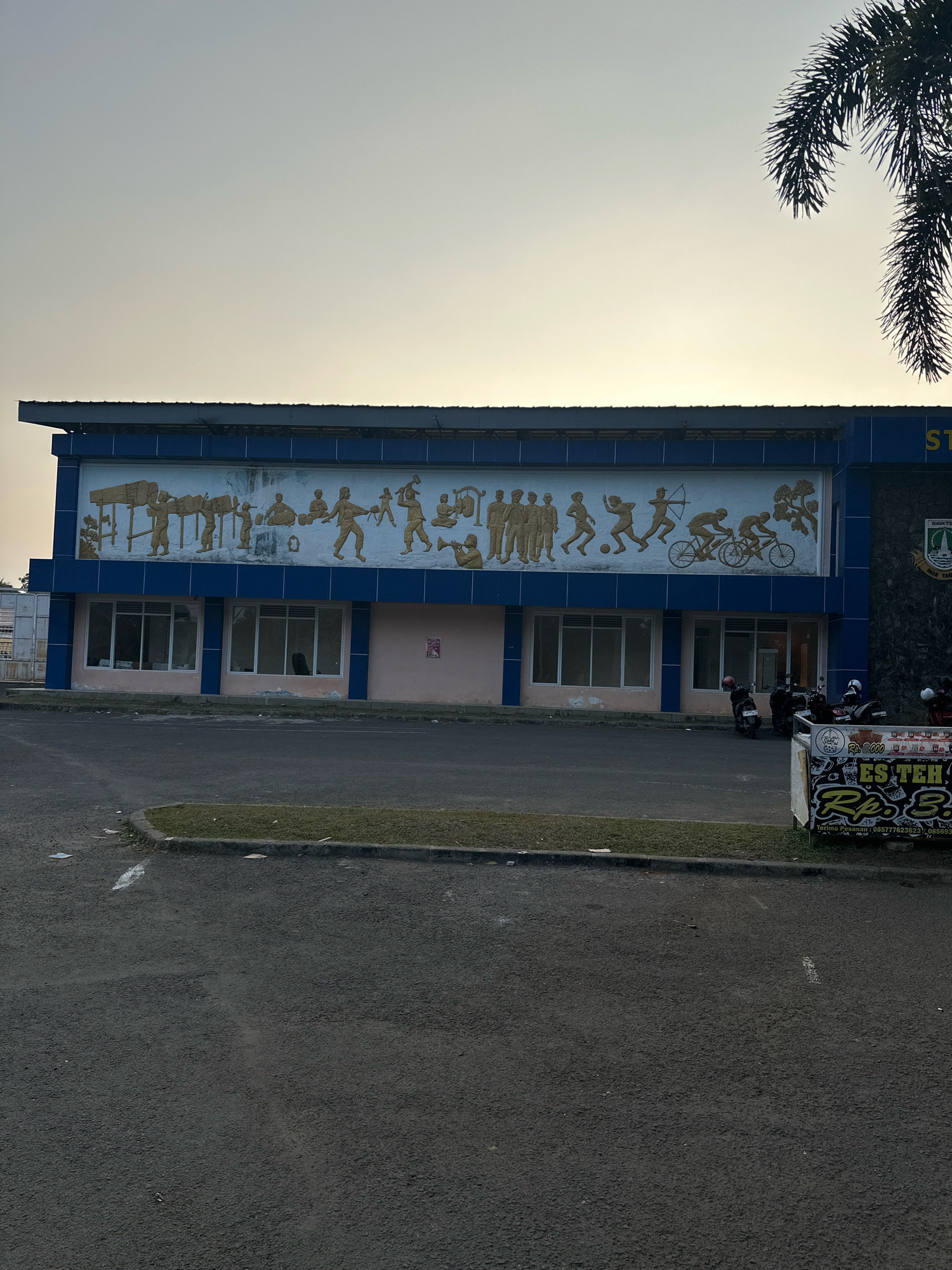The image depicts the exterior of a building with a mural on its facade, located in the city of Saruni, Indonesia. The mural, rendered in a golden hue, portrays various human figures engaged in activities such as farming, athletics, and archery, along with depictions of musical instruments and natural elements. The building itself is painted in blue and pink, with large windows on the ground floor. In front of the building, a paved area serves as a parking lot, with several motorcycles parked. To the right of the building, a sign advertises "ES TEH" (iced tea) with pricing information. The sky is overcast with a pale hue, suggesting either early morning or late afternoon light. A palm frond from a nearby tree is visible in the upper right corner of the frame. The scene is a street view, presenting a slice of urban life in Saruni.