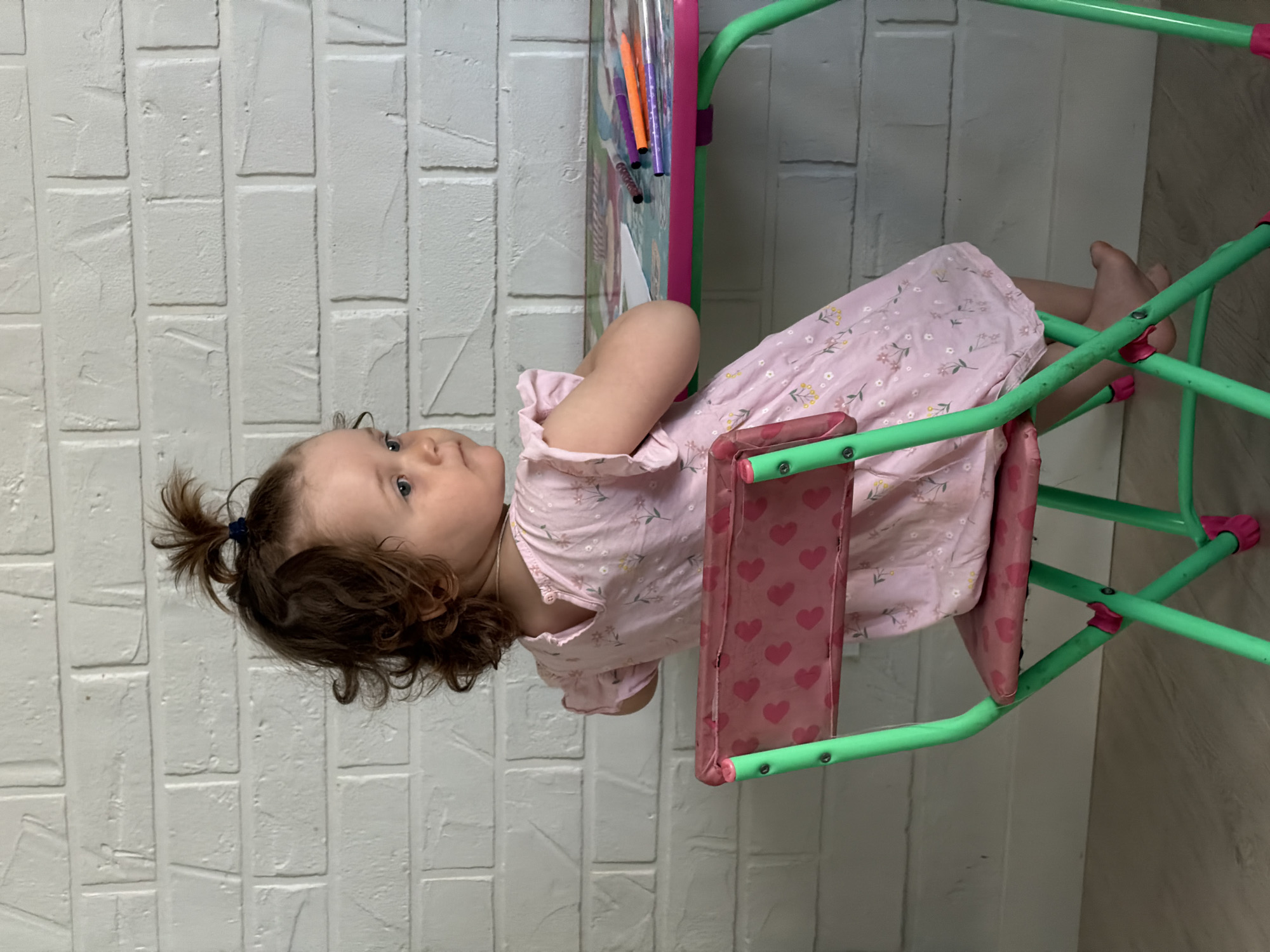 A young child with brown, curly hair pulled up in a small ponytail is sitting on a child's chair and appears to be looking off into the distance. The child is wearing a light pink dress with a floral print. The chair is light green with pink accents on the seat and connecting pieces. There is what appears to be a drawing board on the left side of the child, with colorful markers attached to it. The background is a white brick wall on the left and a light-colored wall on the right. The location is likely indoors in Pervomaysky, Russia.