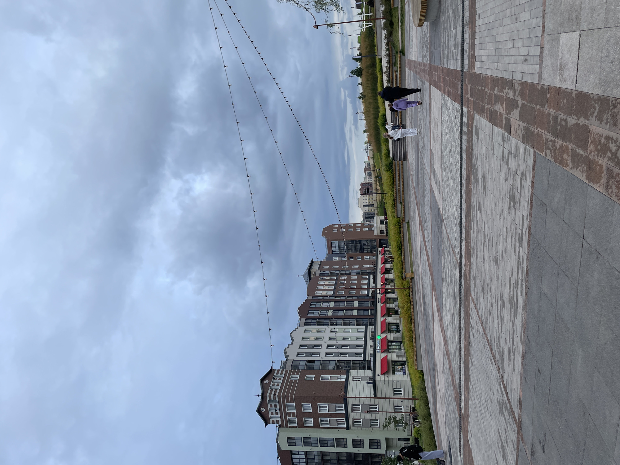 The image depicts an outdoor public space with a paved plaza in the foreground and a multi-story residential building in the background. The sky is overcast with dark clouds. Several strings of decorative lights are suspended across the plaza.
In the plaza, three individuals are walking away from the camera. Two are adults, and one appears to be a child. They are positioned towards the right side of the frame. On the left side of the plaza, a person on a scooter is facing the building.
The building is modern in design, with a varied facade featuring different colors and window styles. Red awnings are visible over some ground-floor storefronts. A row of manicured bushes and some small trees line the front of the building. Beyond the building, a gently sloping landscape with green grass and scattered trees can be seen.
The plaza pavement is made of large, square gray tiles with occasional rectangular brick-like sections. There are also visible drainage grates. Benches are placed along the edges of the plaza. The overall impression is a contemporary urban or suburban development area. The image does not provide specific clues to identify Pervomaysky, Russia, without external information.