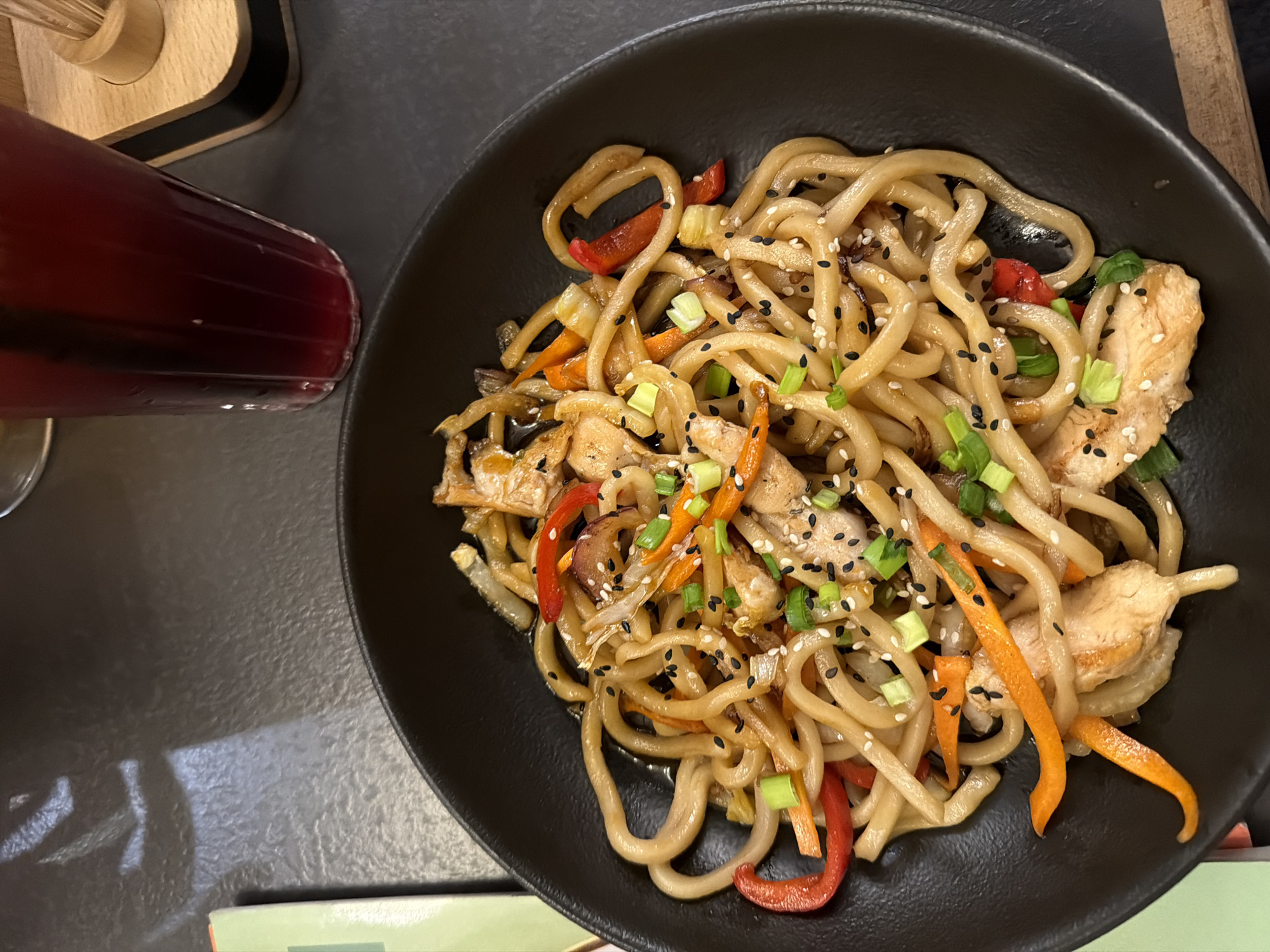 An overhead shot captures a dark, textured table holding a substantial meal, likely in a restaurant or cafe setting in Saint Petersburg, Russia. The central focus is a large, round black bowl filled with udon noodles. The noodles are mixed with sliced chicken breast, vibrant orange julienned carrots, strips of red bell pepper, and what appear to be thinly sliced onions or other vegetables. The dish is generously garnished with chopped green onions and a sprinkle of both white and black sesame seeds, suggesting an Asian-inspired cuisine. The noodles and other ingredients are coated in a glossy, light brown sauce.
To the left of the bowl, a tall glass contains a dark red or purple beverage, possibly juice or a berry drink. In the upper left corner, a wooden holder with several thin sticks, likely chopsticks or toothpicks, is partially visible. A sliver of a light green object, possibly a menu or book, can be seen along the bottom edge of the frame. The table surface is dark and appears slightly reflective, with light sources creating subtle glares. The overall impression is one of a casual dining experience. No discernible text is visible in the image.