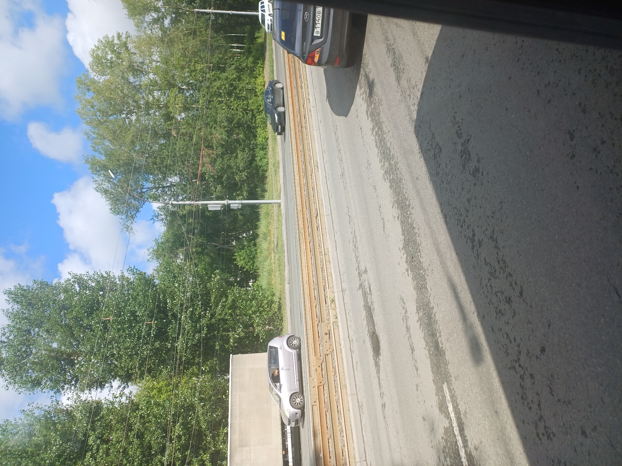 In Novosibirsk, Russia, on a sunny day with a partially cloudy blue sky, a scene unfolds on a multi-lane road adjacent to tram tracks. In the foreground, the asphalt surface shows patches of wetness and is partially obscured by a shadow cast from the right side of the frame, likely from a vehicle or structure.
On the road, a dark gray sedan, identified by its license plate "B114OB1," is visible from the rear, moving away from the viewer. Further ahead, a dark gray sedan is also traveling in the same direction. To the left, a light silver hatchback is seen from its side, with a person visible inside. Behind the silver car, the side of a light-colored box truck is also present. In the distance, a white van is observed.
The environment is characterized by a line of lush green trees forming a dense backdrop to the road. Overhead, power lines are strung between poles, and a street light is positioned to the left of the frame. The tram tracks run parallel to the road, separated by a raised, weathered embankment.