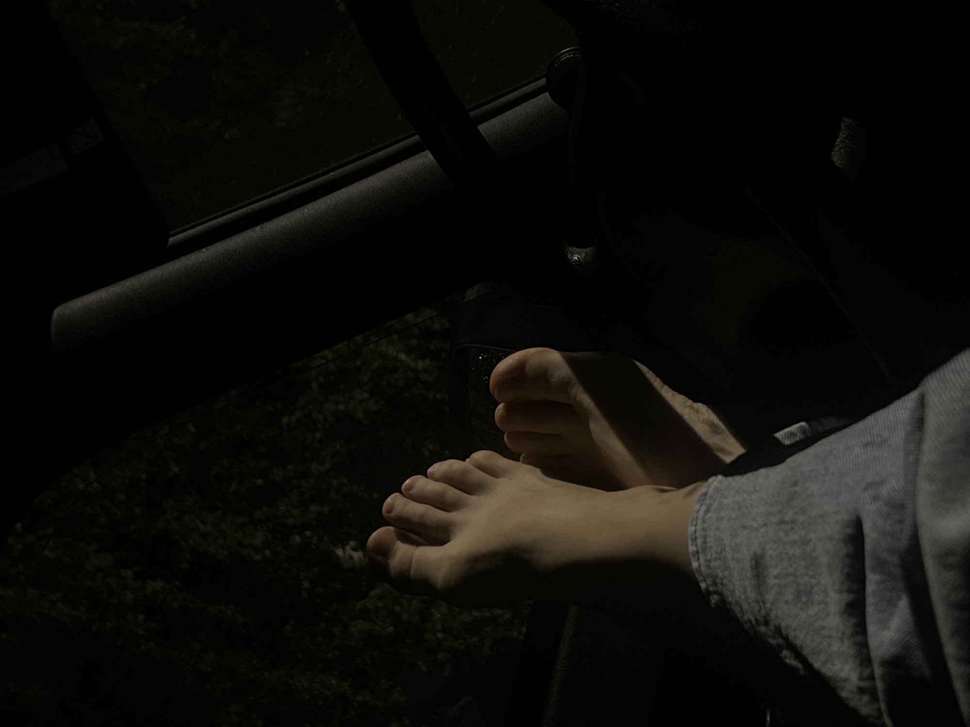 In Staroe Mart'yanovo, Russia, a close-up shot reveals a person's bare feet and lower legs resting inside a vehicle at night. The feet are illuminated by an unseen light source, casting shadows. The individual is wearing light blue jeans. The background outside the vehicle is dark and appears to be foliage, suggesting a rural or wooded location. The interior of the car is dimly lit, with the focus on the feet and the immediate surroundings.
